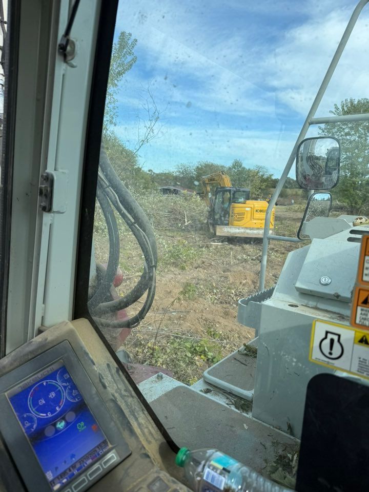 View from machinery cab, overlooking clearing with a yellow excavator in the background under a blue sky.