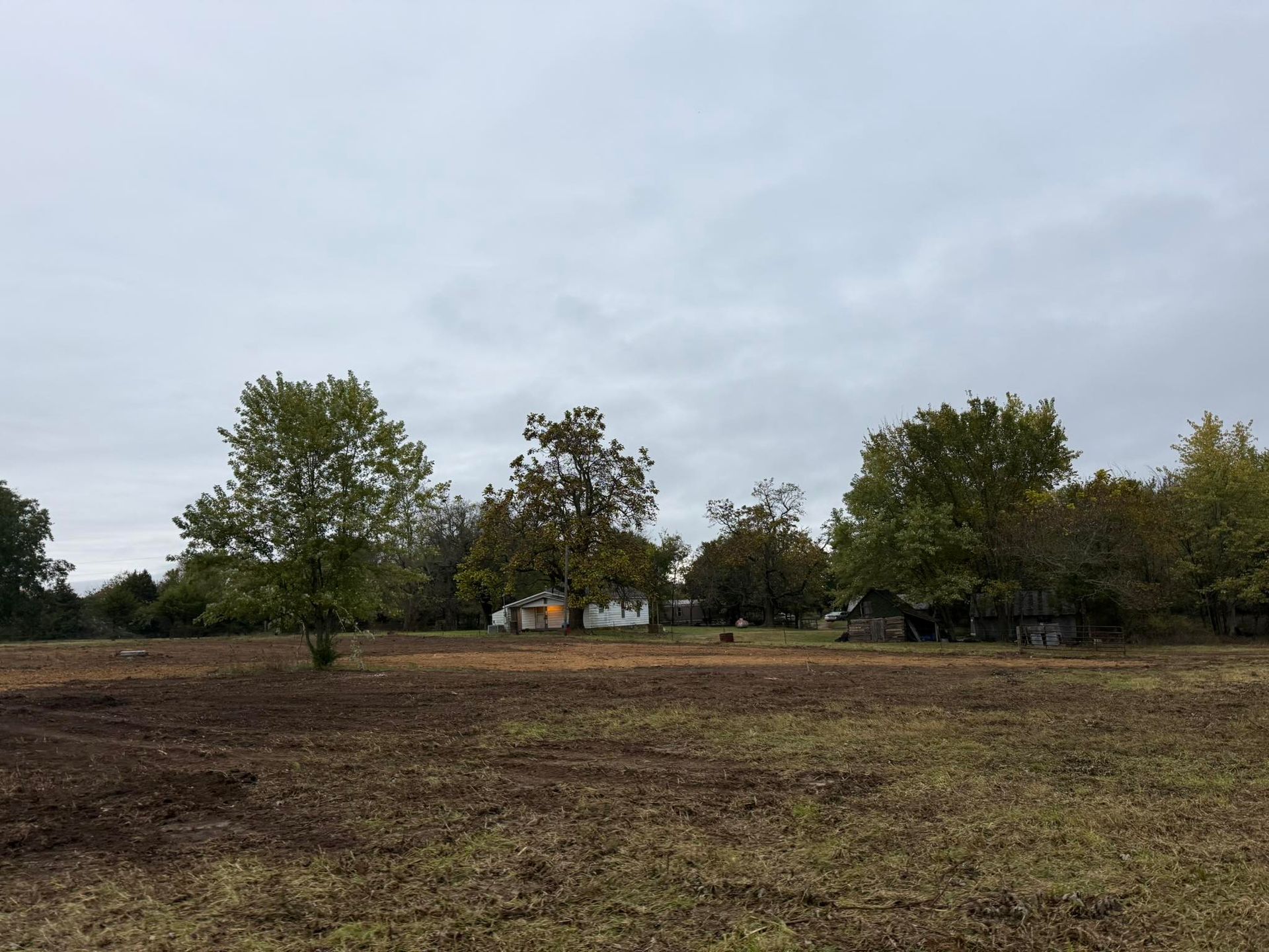 Field with bare earth, trees, and small, weathered buildings under a gray, overcast sky.