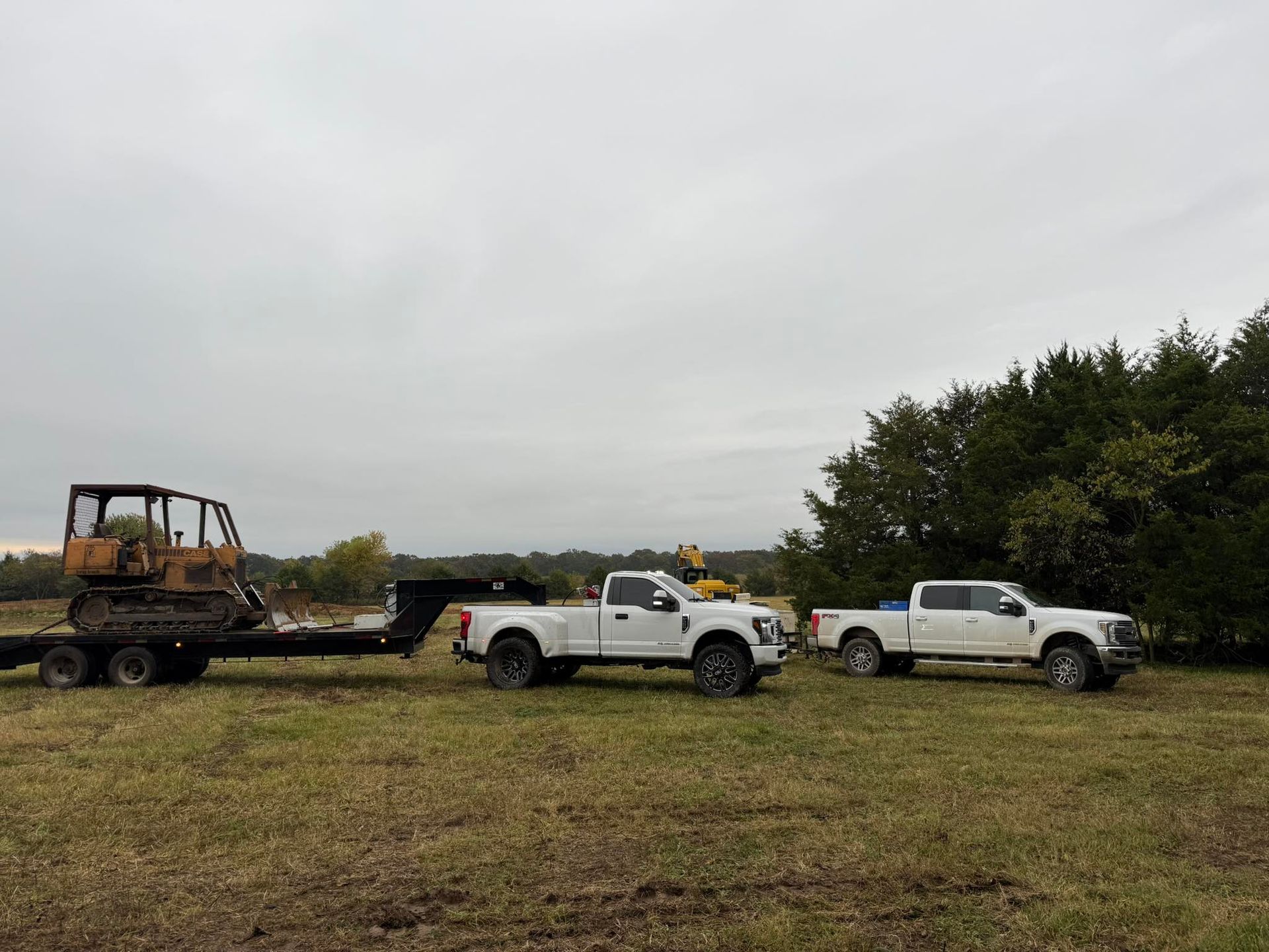 Two white pickup trucks, one towing a trailer with a small bulldozer, in a grassy field under a cloudy sky.