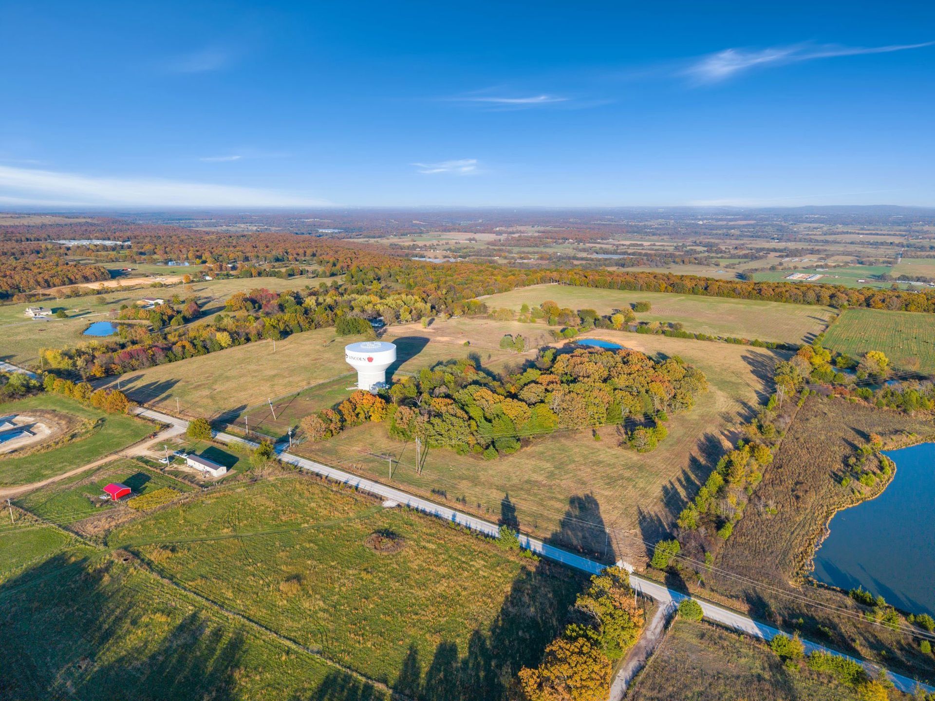 Aerial view of rural landscape with water tower, fields, ponds, and trees under a blue sky.