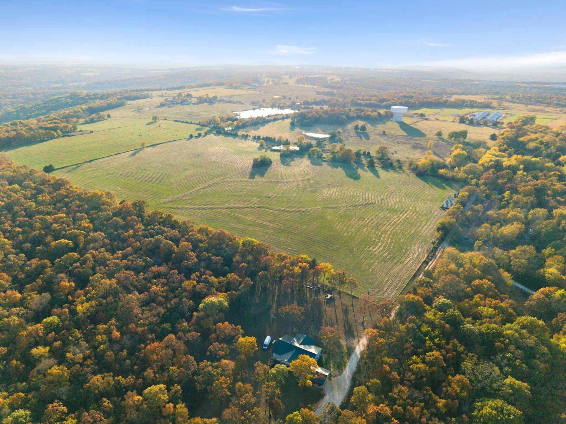 Aerial view of a rural landscape with a house, fields, trees, and water tower under a blue sky.