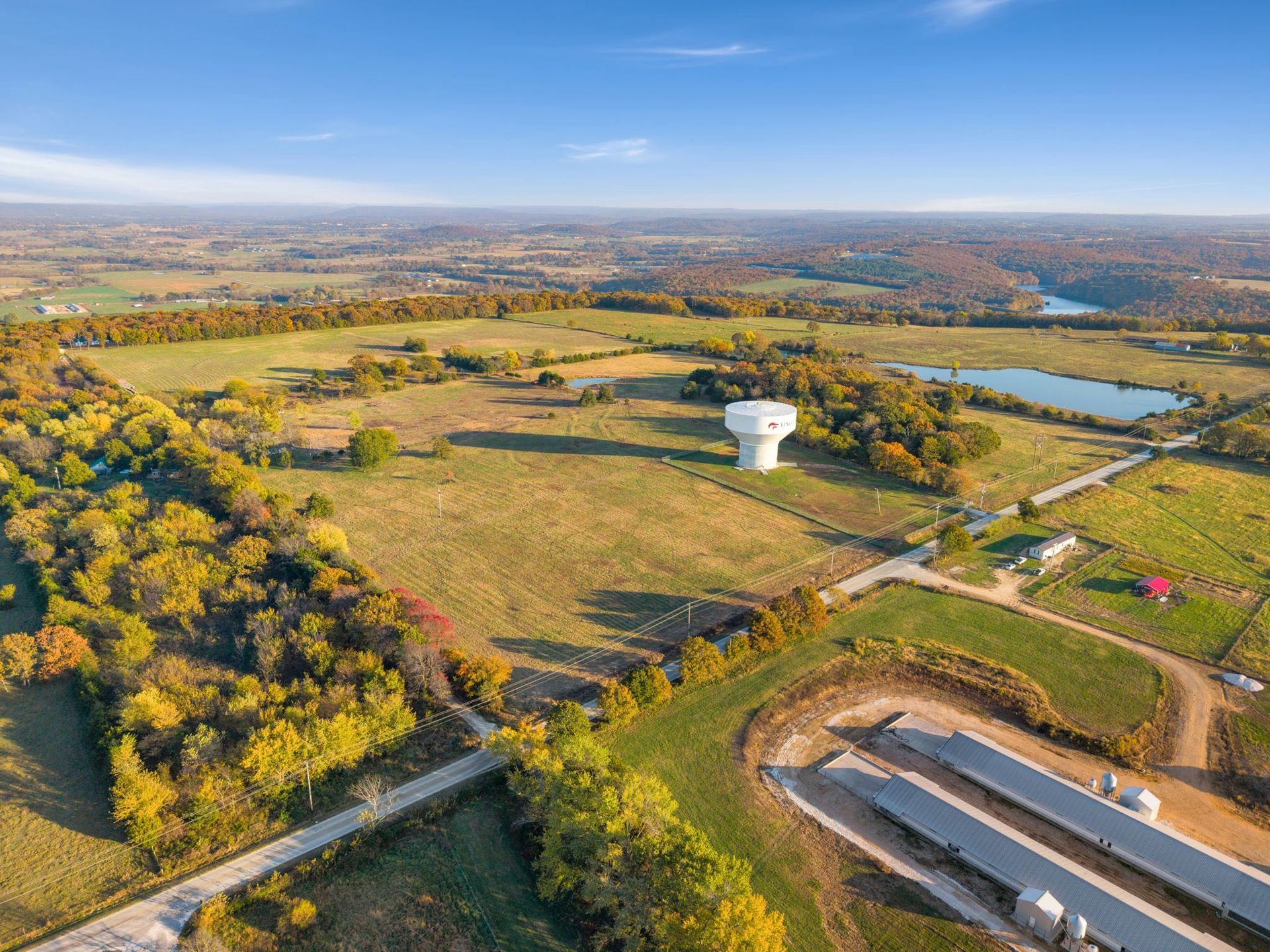 Aerial view of a rural landscape with a water tower, fields, trees, and a pond under a blue sky.