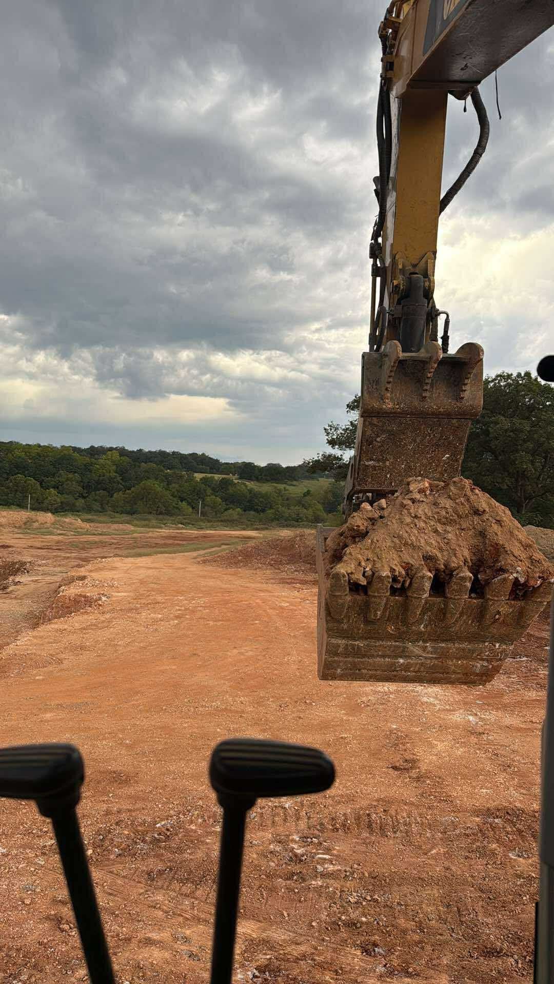 An excavator bucket filled with dirt, in a construction site with cloudy sky.