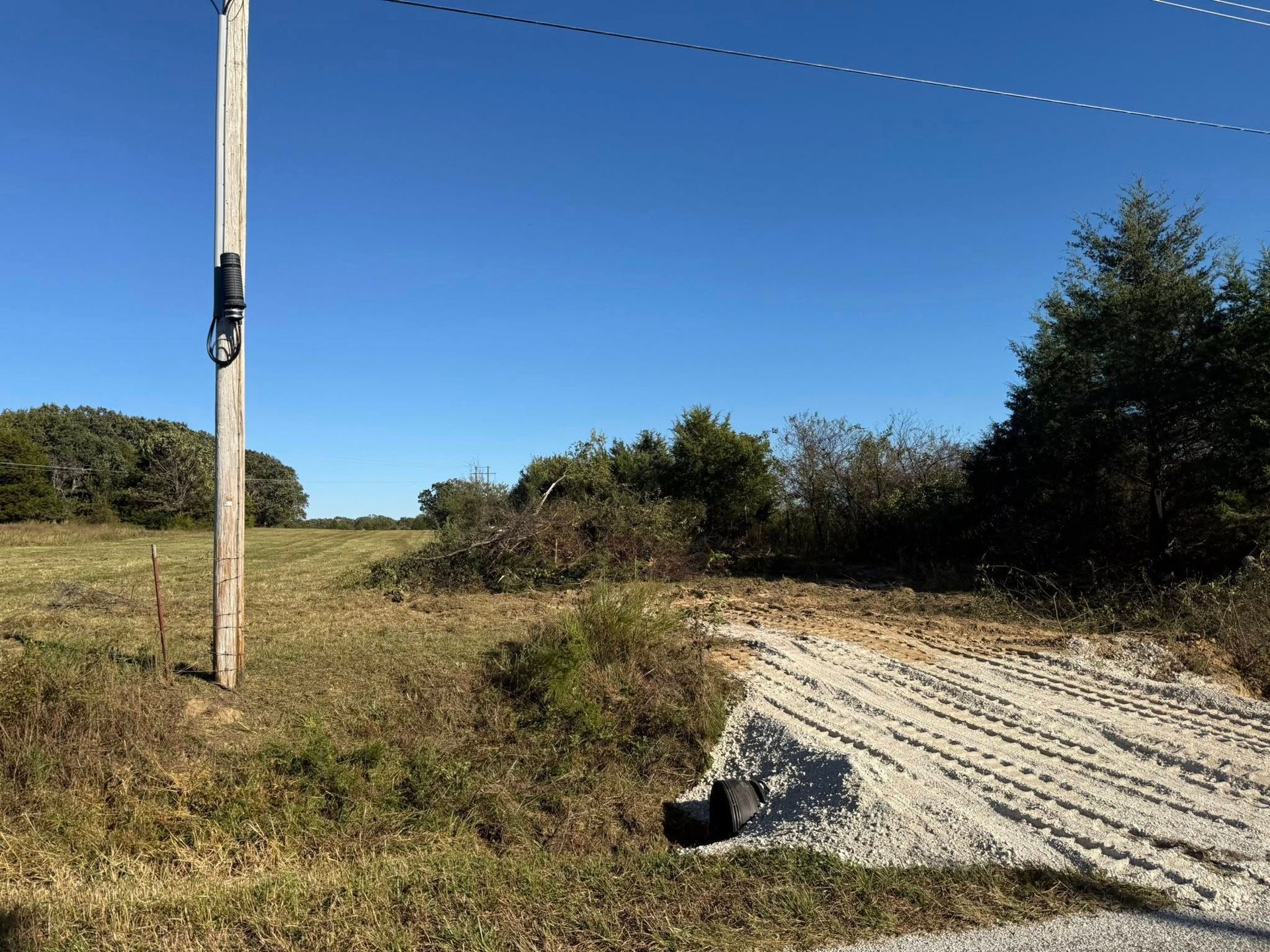 Grassy field next to a utility pole under a blue sky, with a gravel driveway and brush in the background.