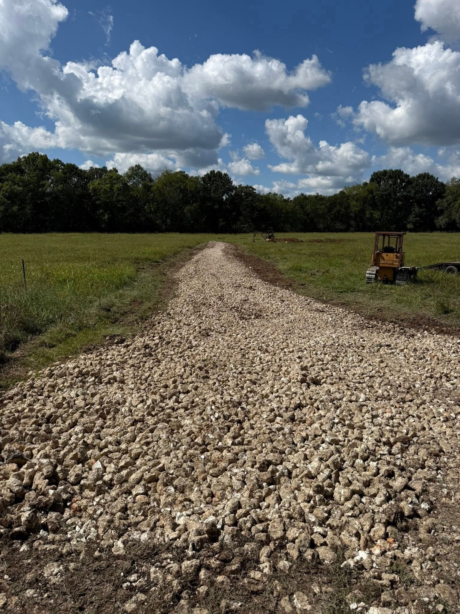A gravel path through a green field leads to trees under a blue sky with clouds.