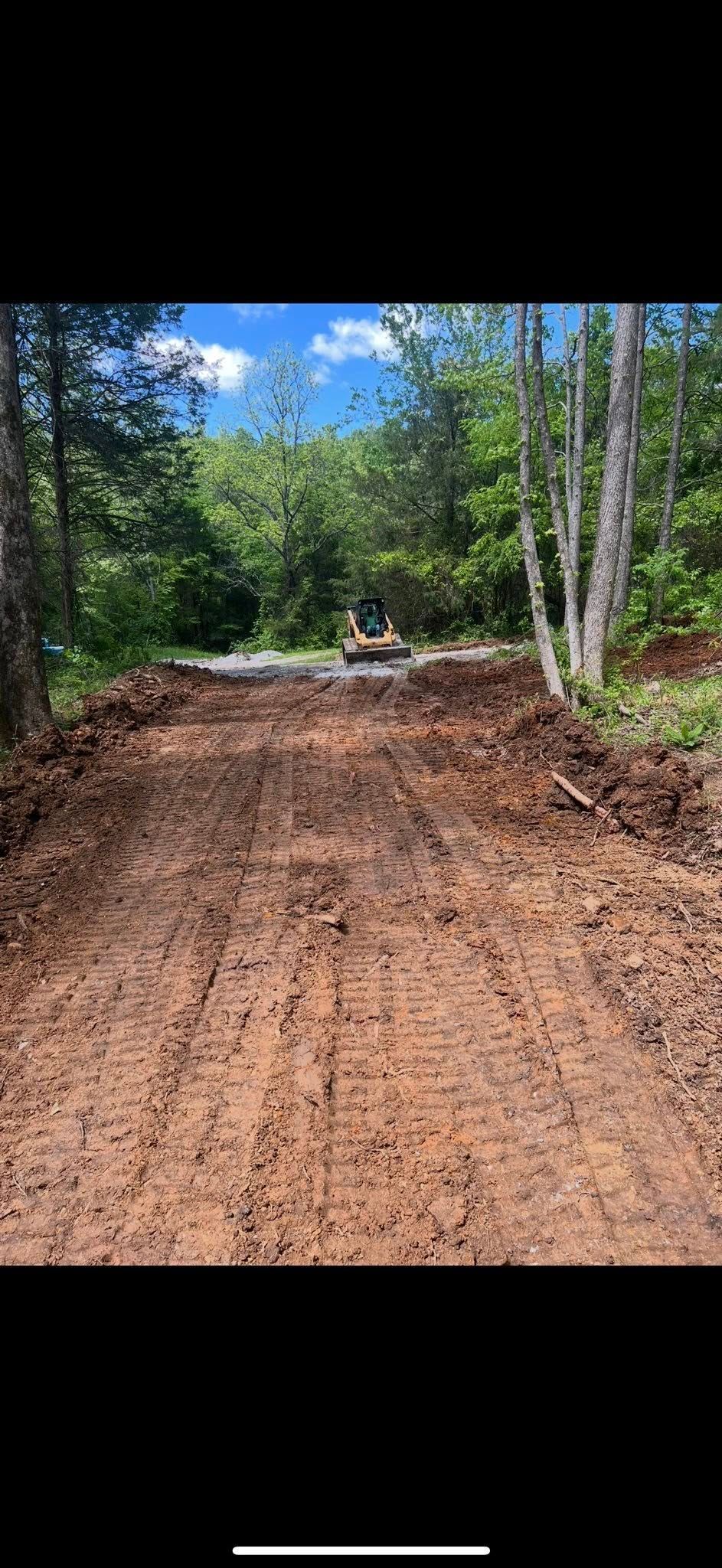 A muddy dirt path through a forest. A small machine is visible at the path's end. Trees and a blue sky are behind.
