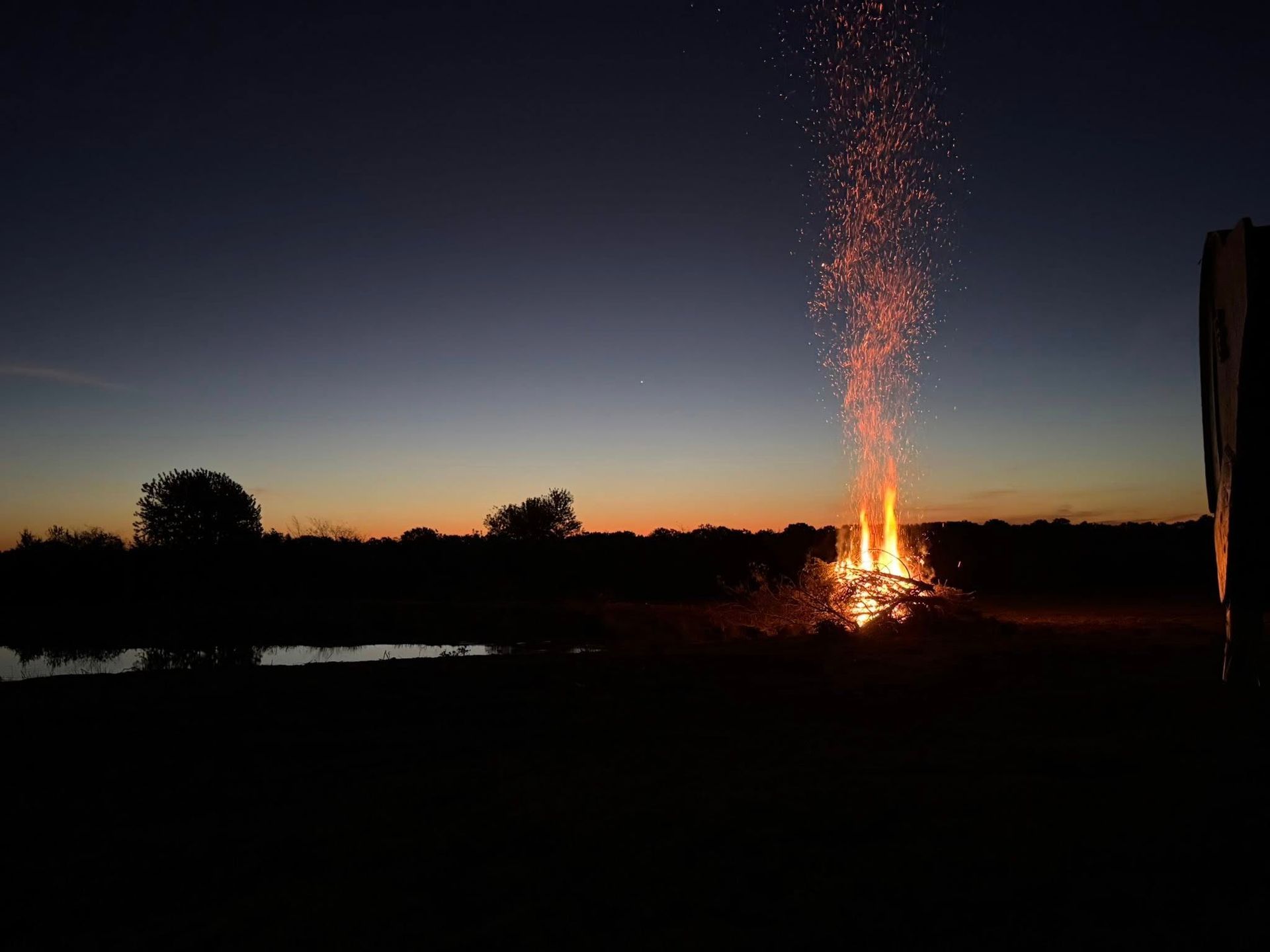 Large bonfire erupts sparks into the twilight sky over a dark landscape, possibly a field near water.