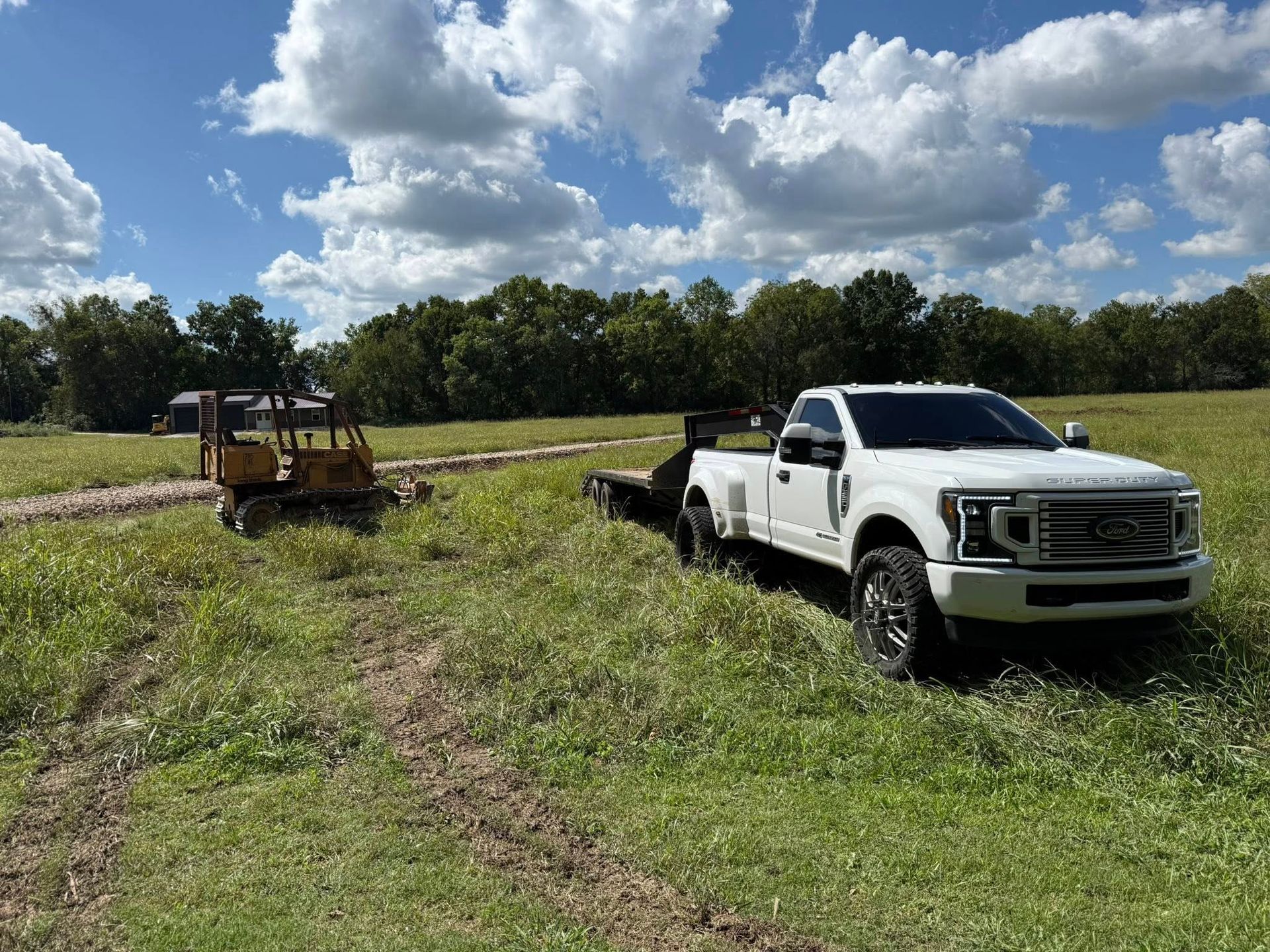 White pickup truck towing a trailer parked in a field with a small yellow machine. Cloudy sky.