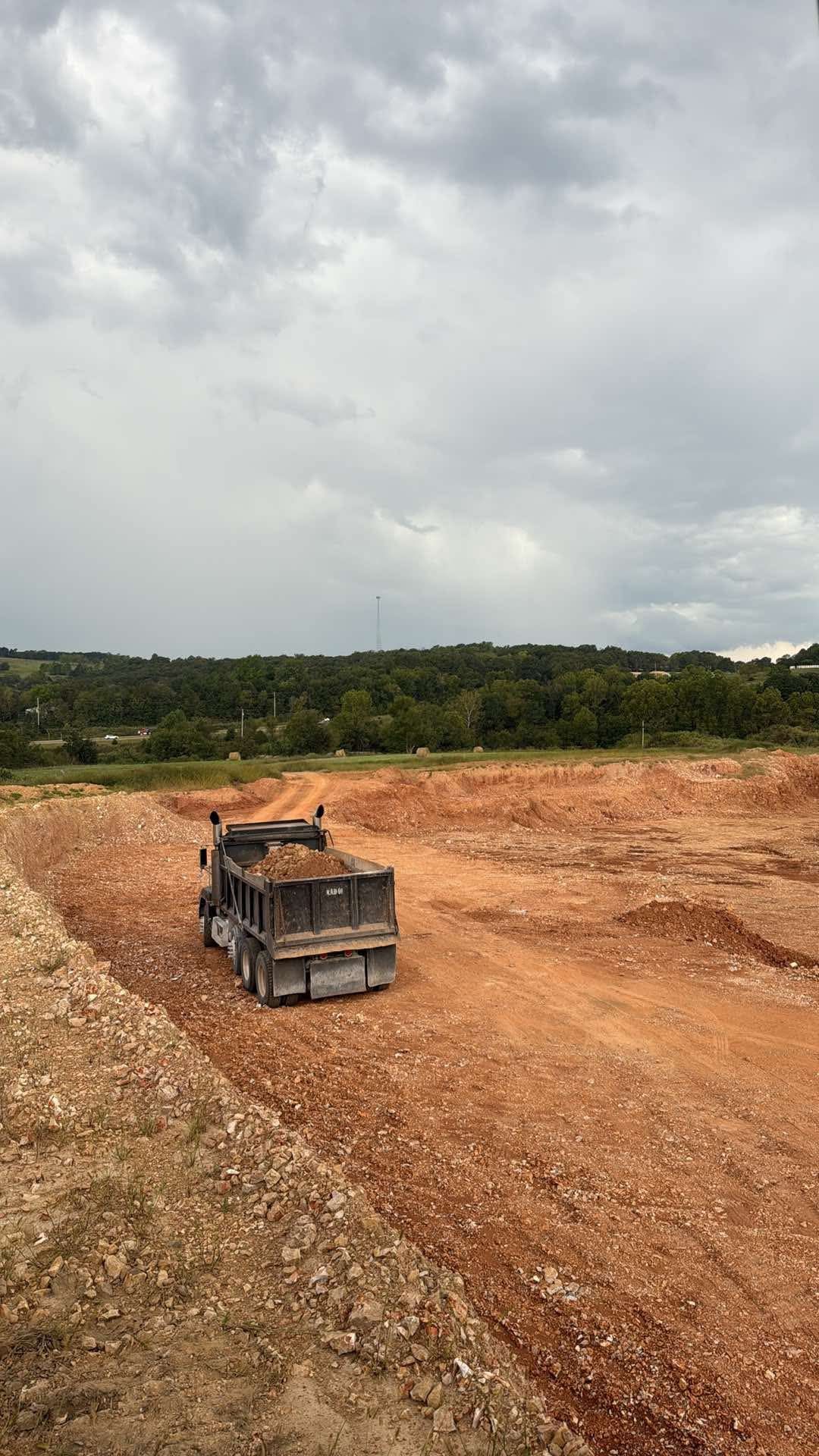 Dump truck on a reddish-brown dirt road, hauling a load, under a cloudy sky near green trees.