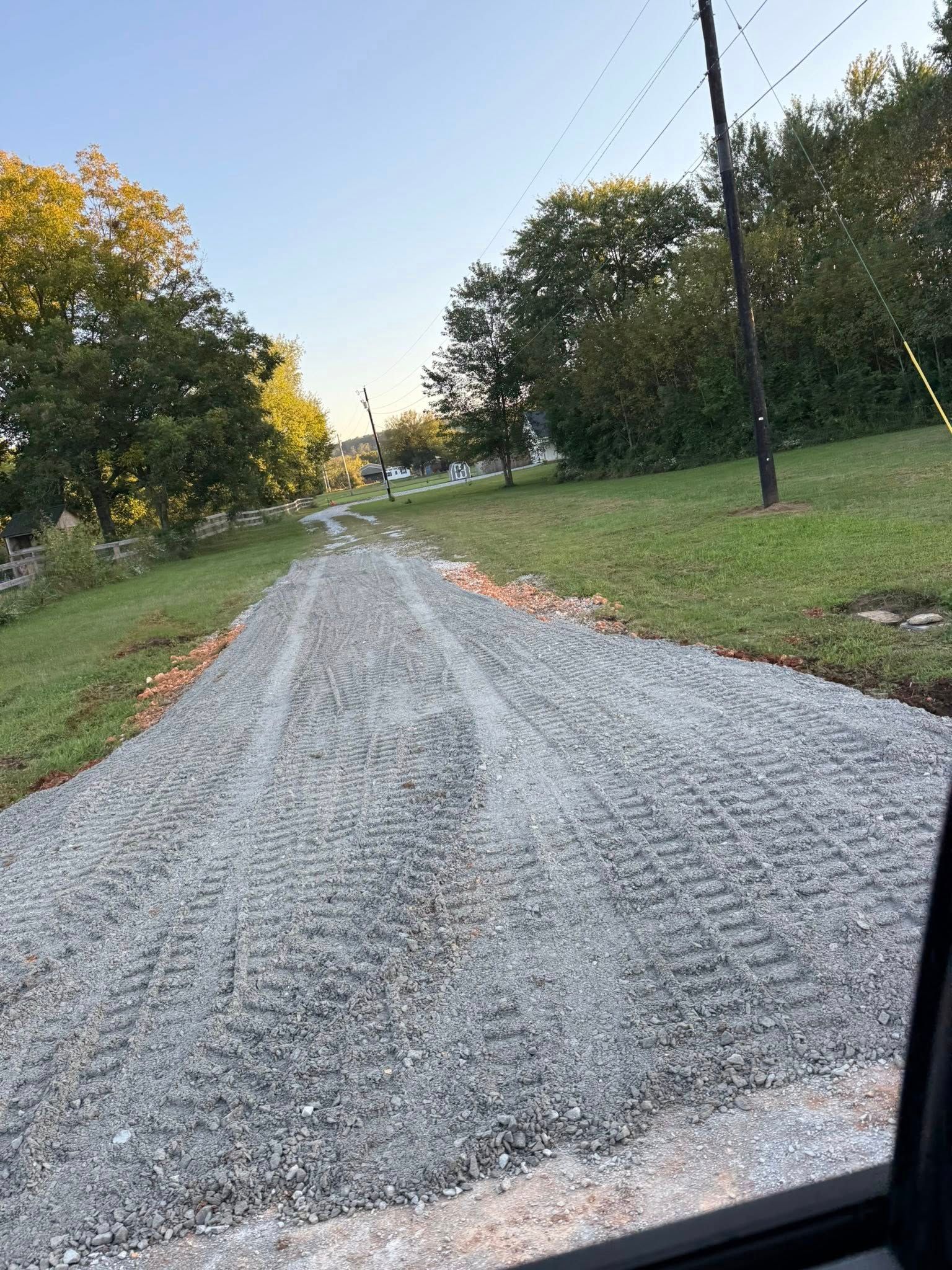 A gravel driveway with tire tracks leads up a slight hill, flanked by grass and trees.