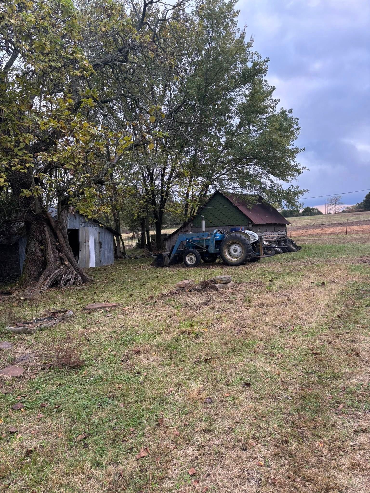 A blue tractor parked near small sheds and trees in a grassy field under a cloudy sky.