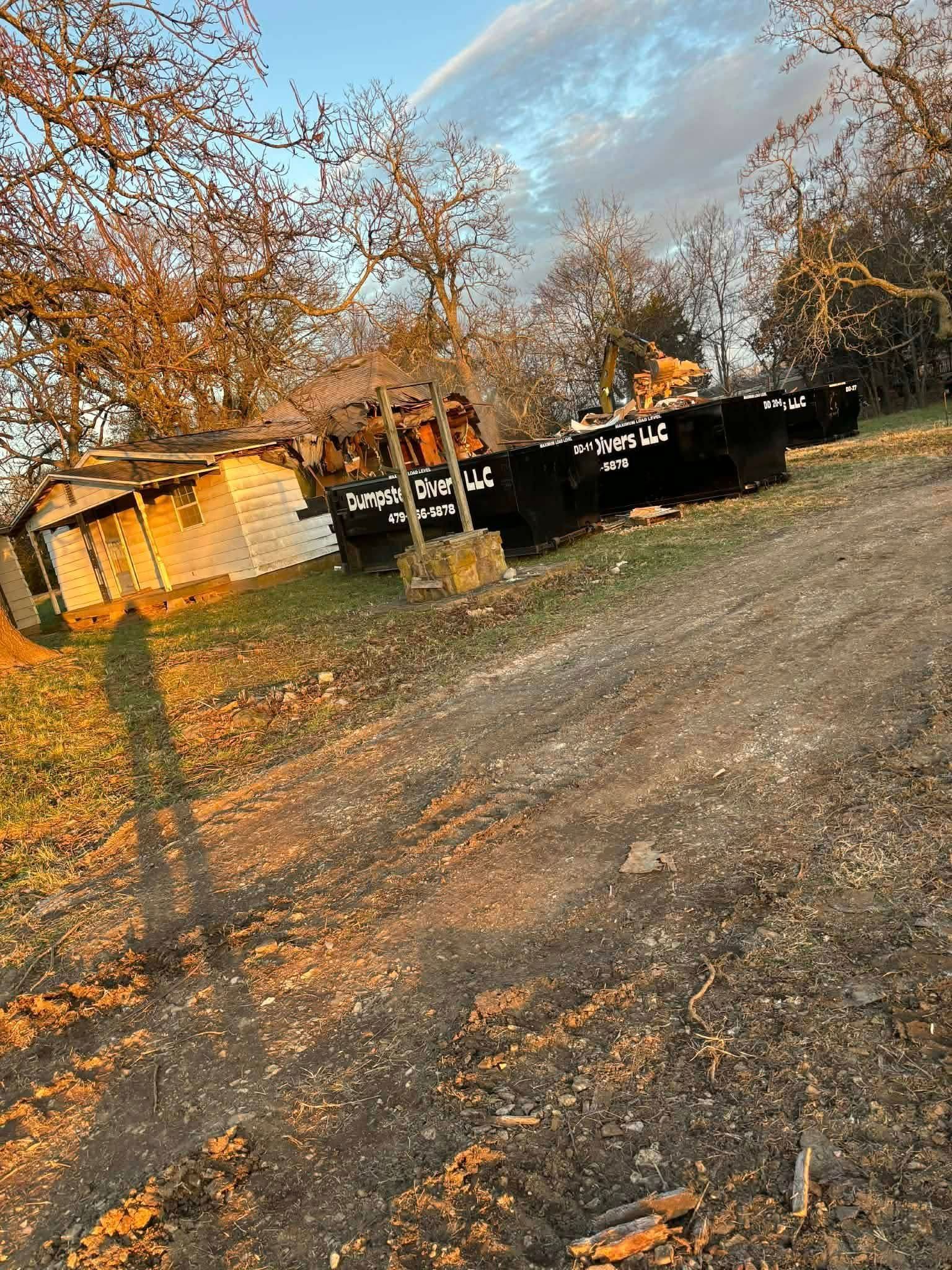 Dirt road with a building and dark trailer under a cloudy sky. Trees and grass in the surrounding area.