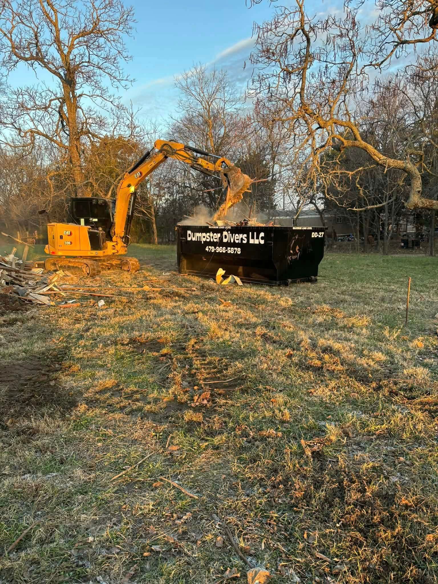 Yellow excavator dumping debris into black dumpster on grassy land. Bare trees in background.