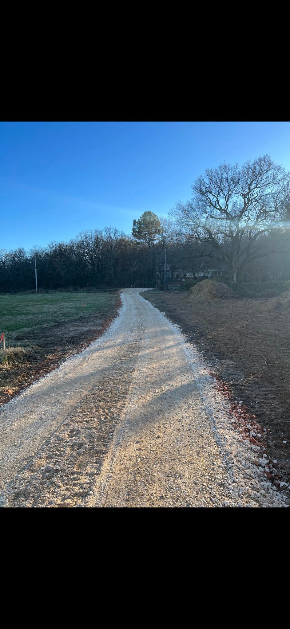 A gravel road leads through a grassy area towards a line of trees under a blue sky.