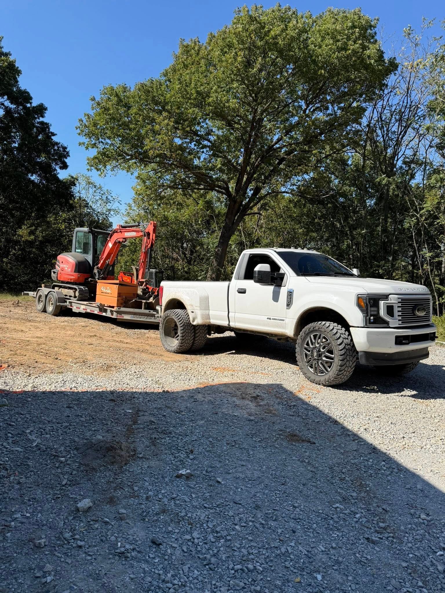 White pickup truck towing a small orange excavator on a trailer on a gravel lot near trees on a sunny day.