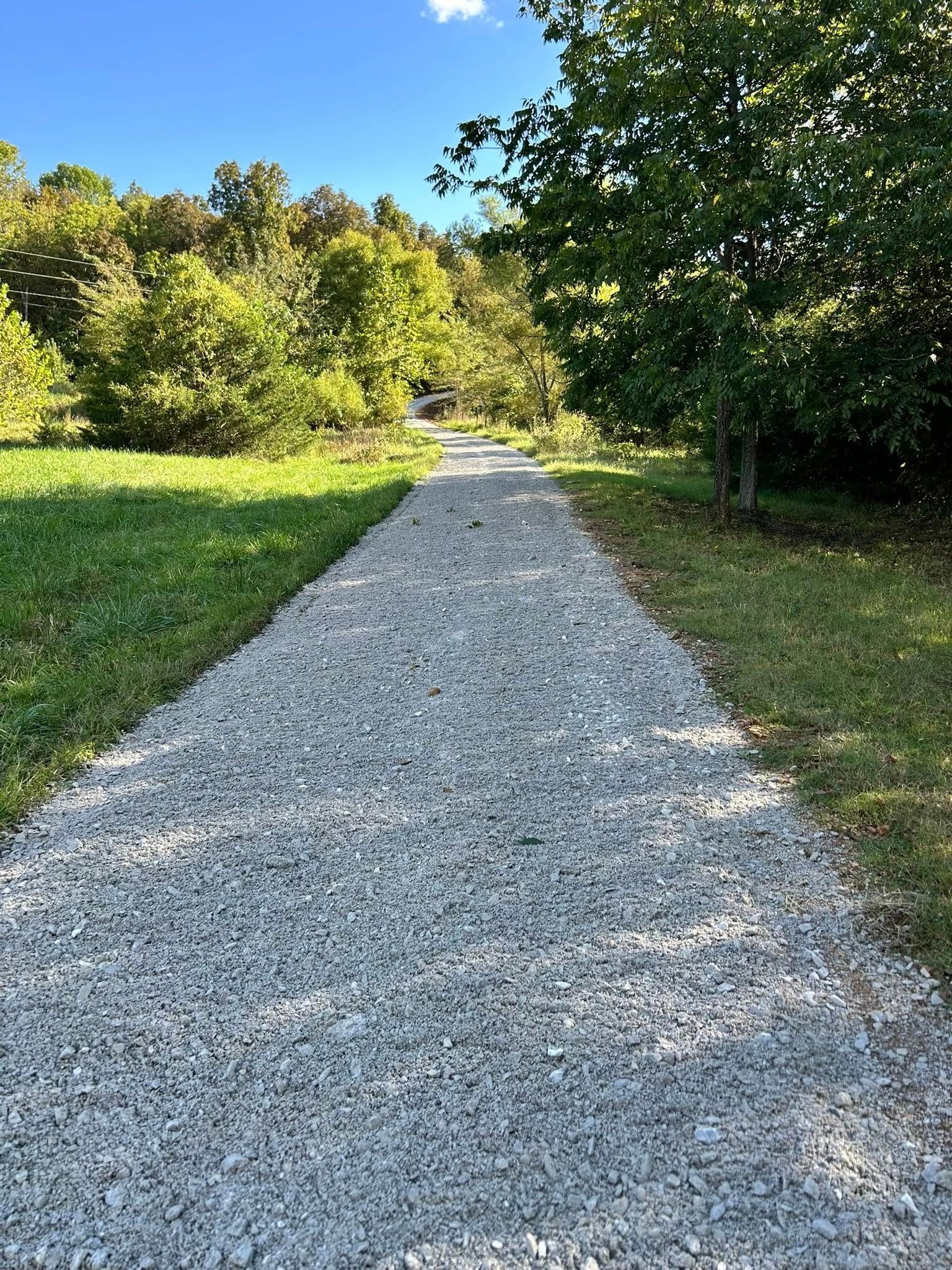 Gravel path through a grassy area, leading into a wooded area on a sunny day.