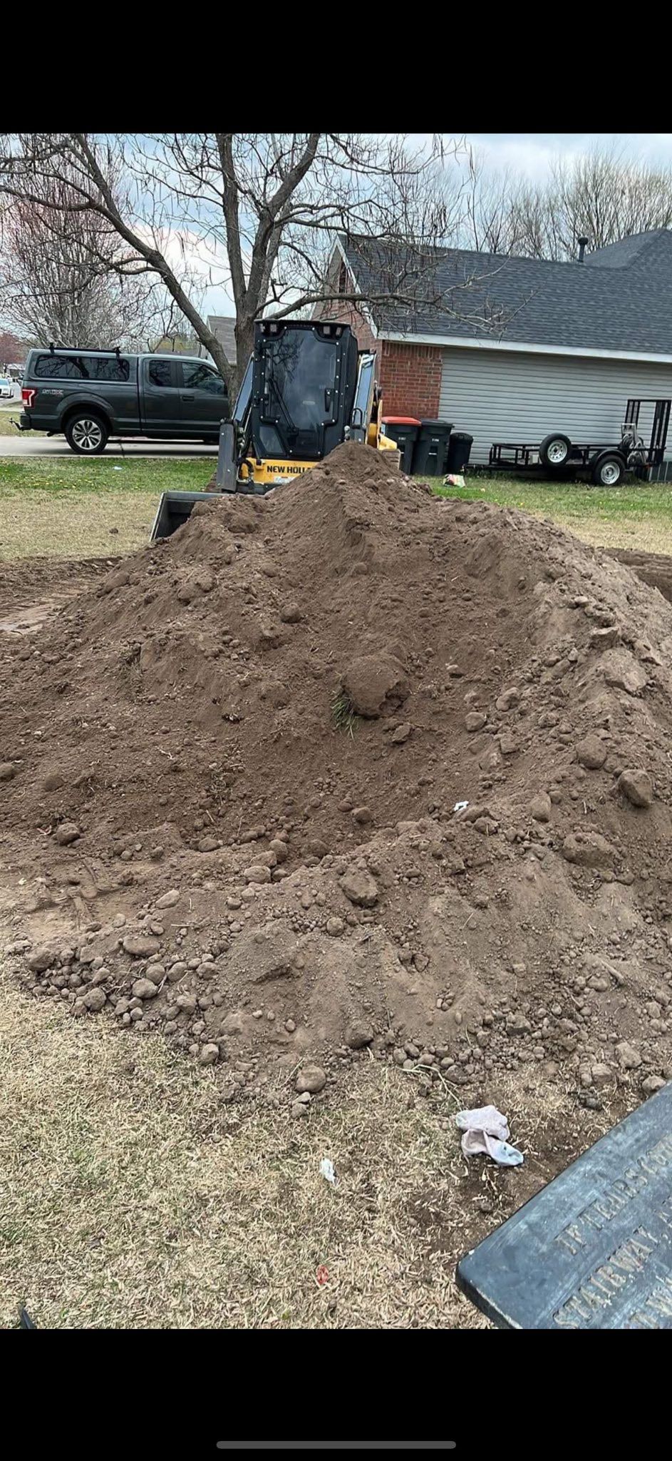 A large pile of dirt in a yard, with a tree and vehicles visible in the background.