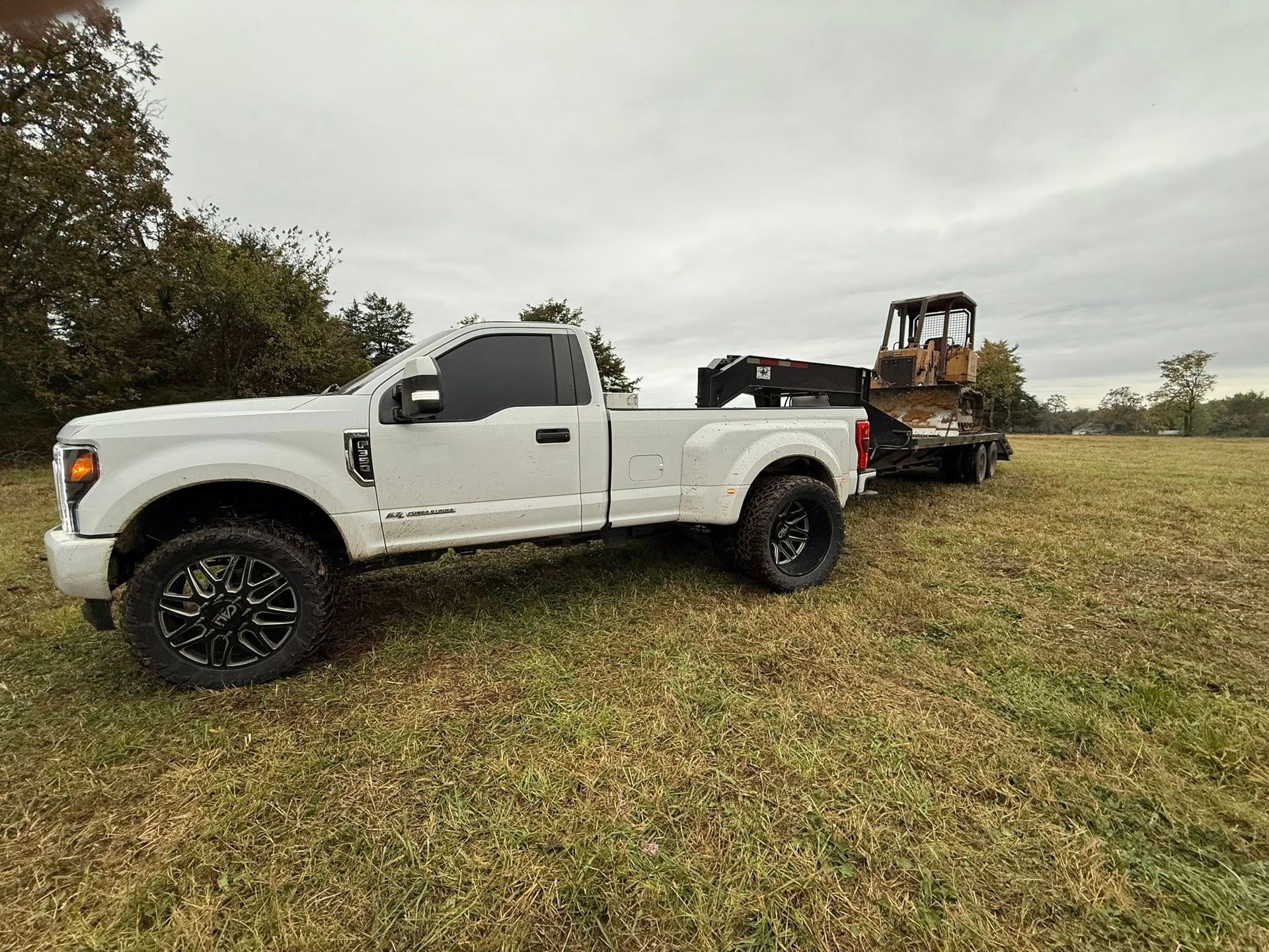 White pickup truck towing a trailer with construction equipment on a grassy field under an overcast sky.