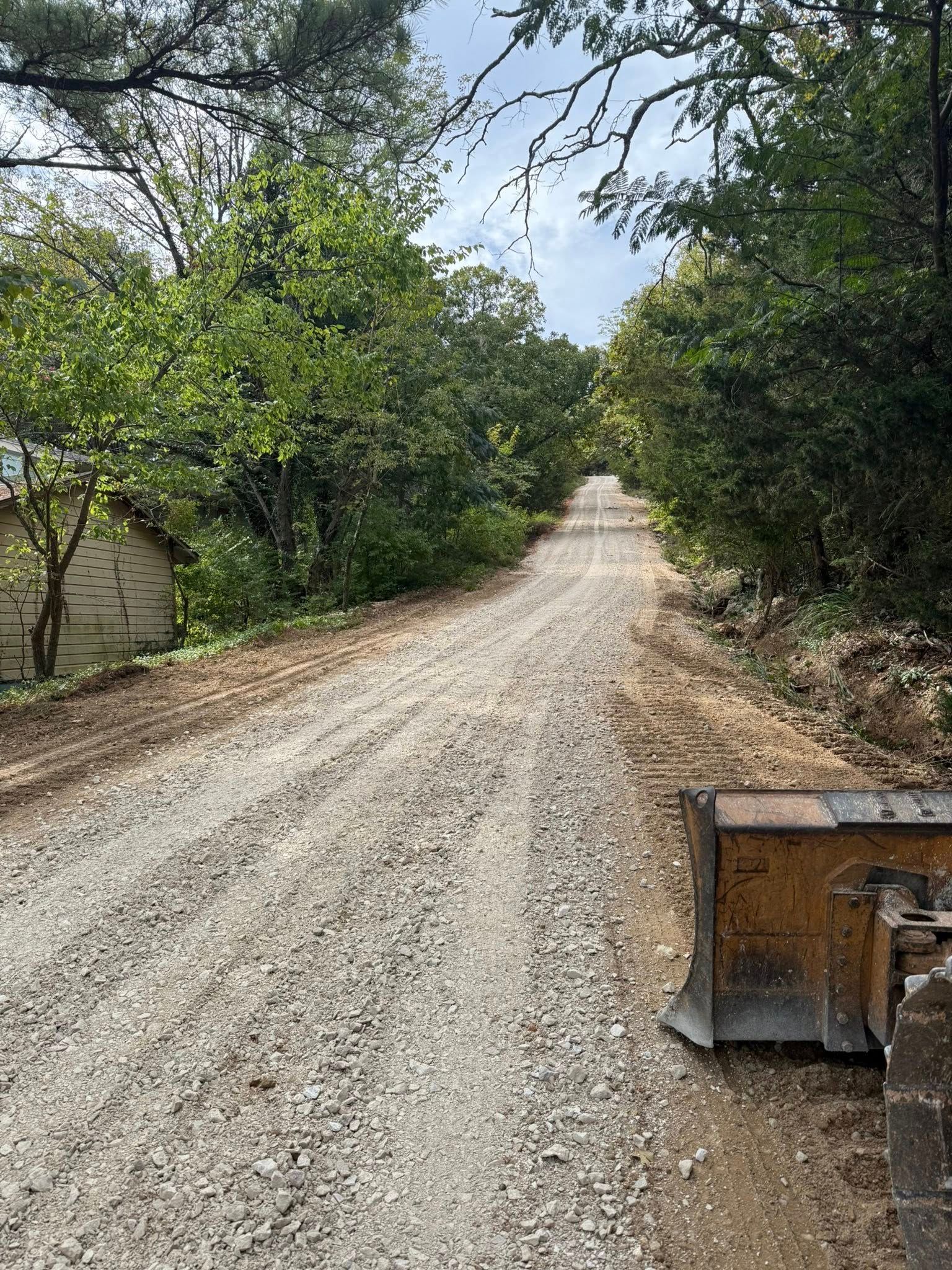 Gravel road through a wooded area. A tractor blade is in the lower right corner.