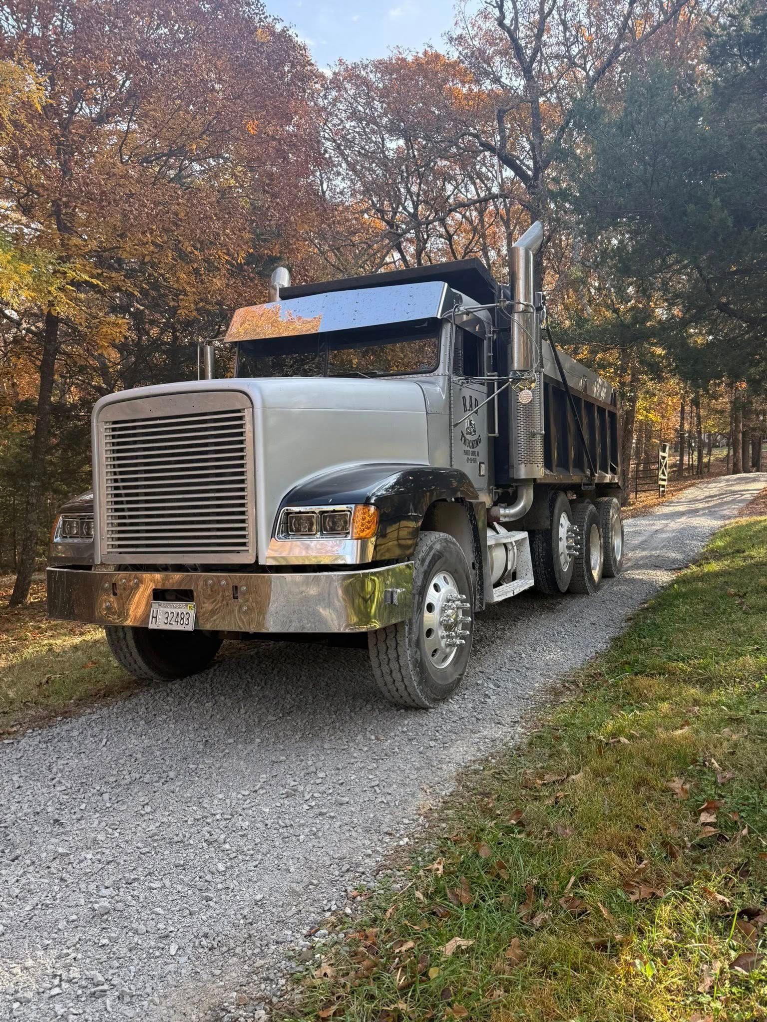 Silver dump truck on a gravel road, autumn trees in the background.