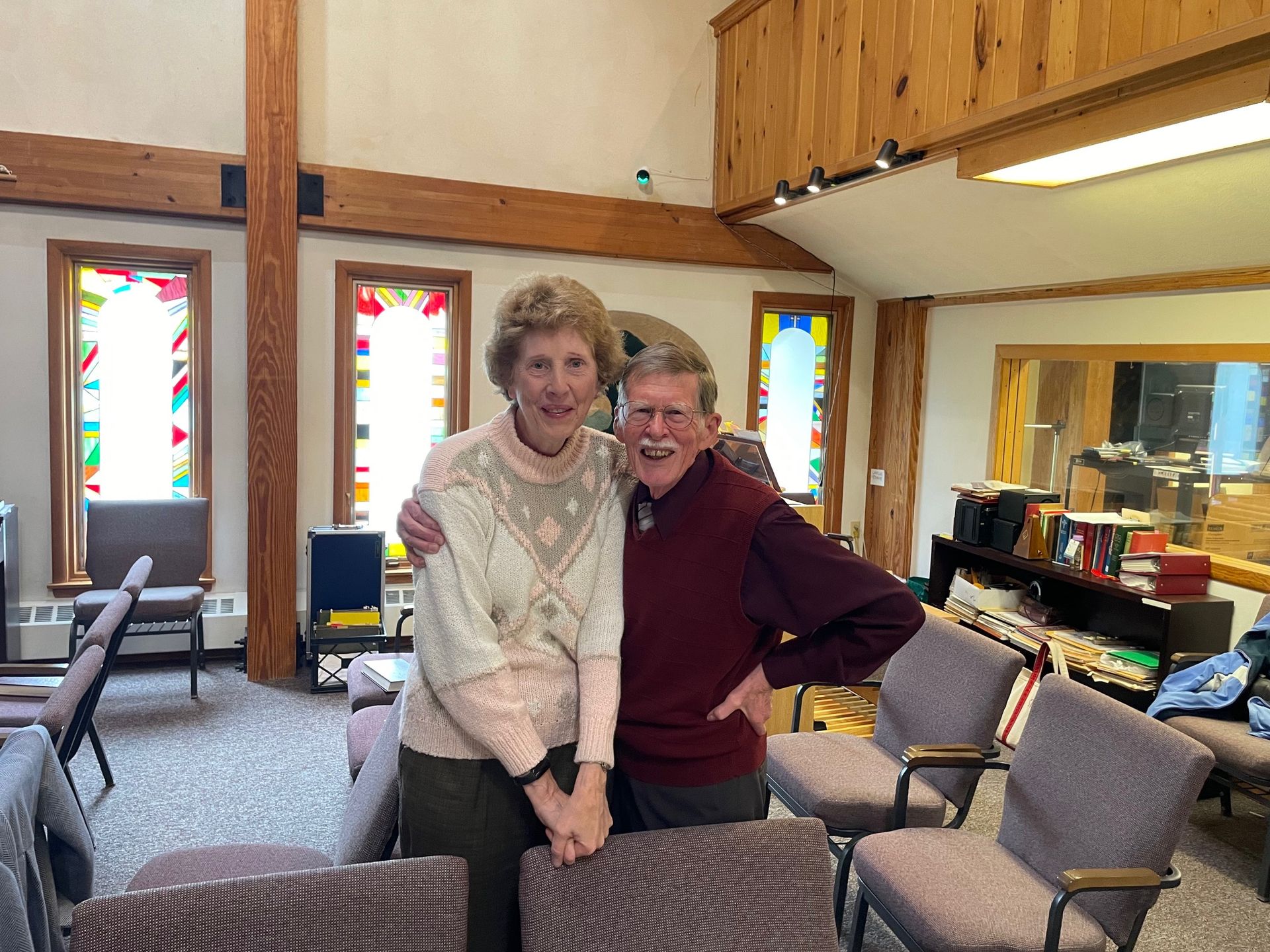 A man and a woman are posing for a picture in a room with chairs.