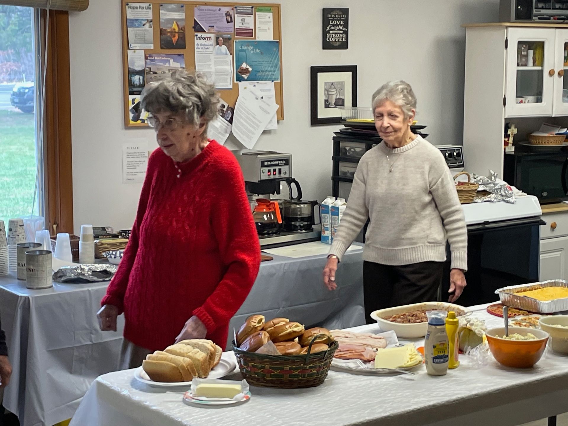 Two older women are standing in front of a table with food on it.