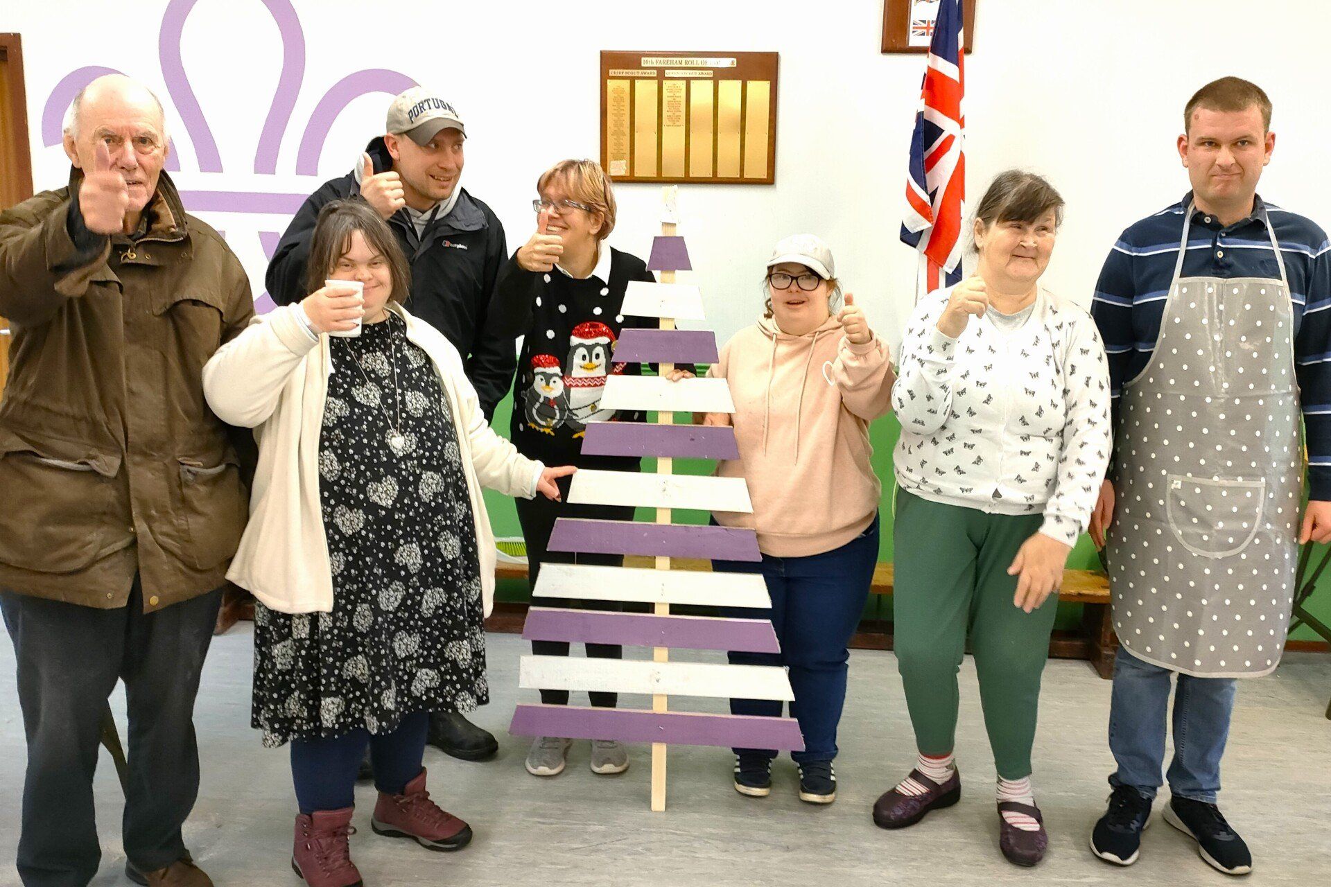 Several FNC members stand and smile, most with thumbs up, holding 1.5 metre  tall  finished banded alternate purple and white painted wood nailed to central piece forming a decorative triangular tree shape.