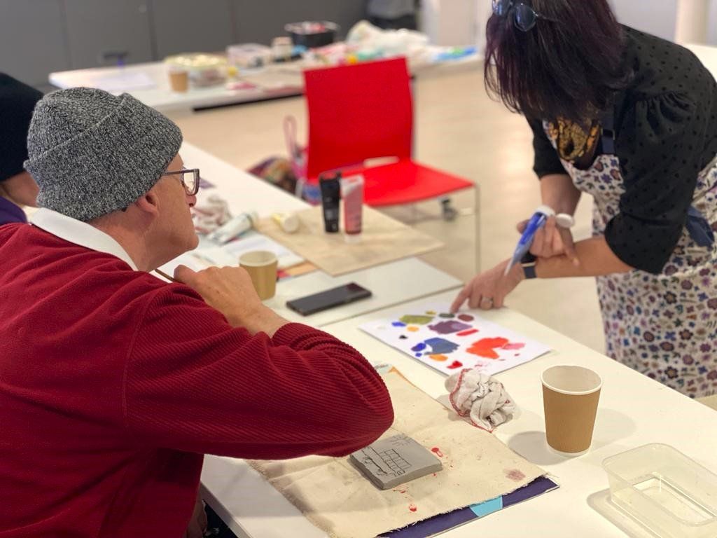 Male FNC Member sat at table with clay tile with a vehicle and Sun carved into it and listens  to a volunteer  who is holding a blue tube of colouring and pointing at different blobs of colour on a piece of paper
