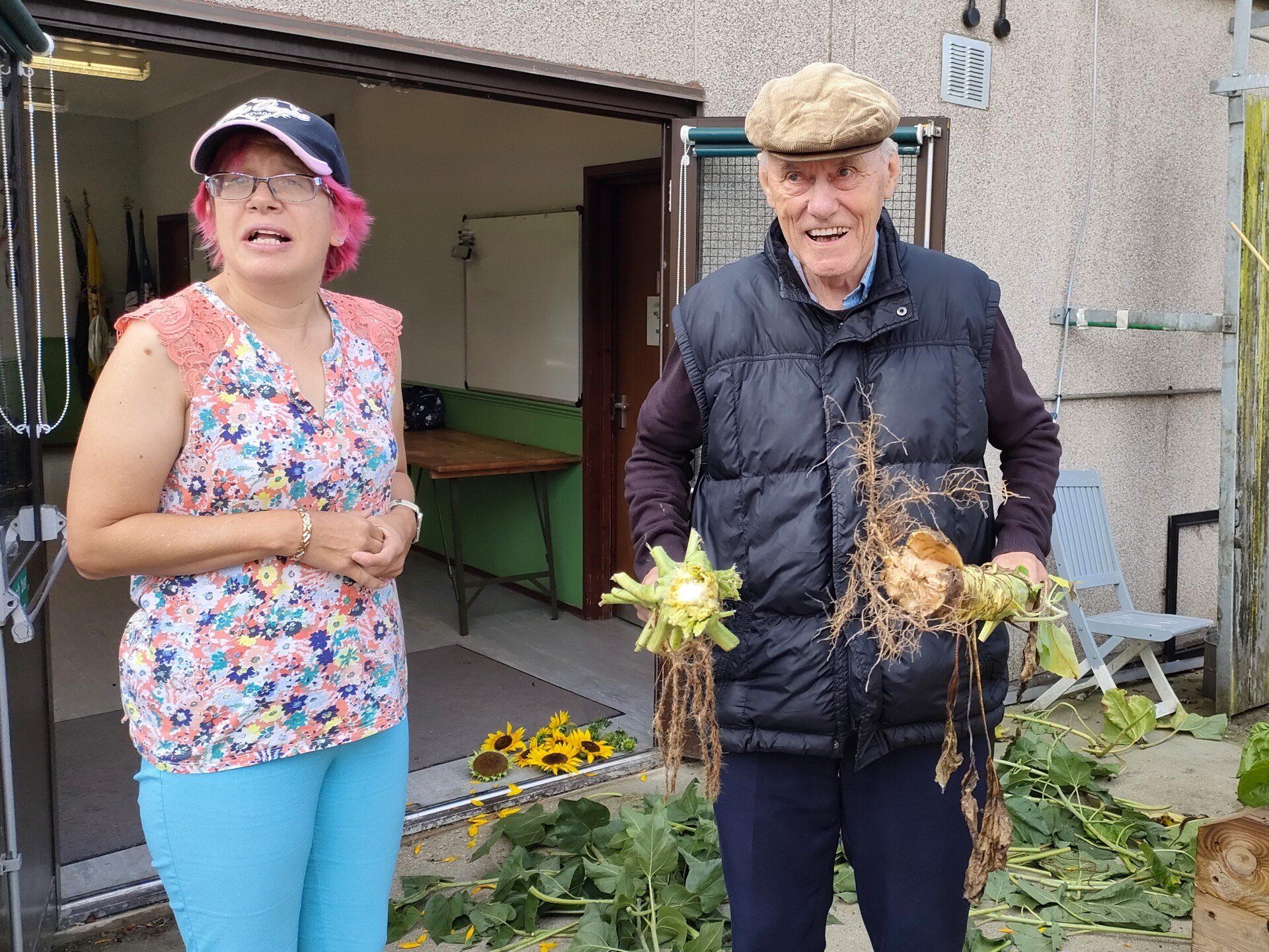 Two smiling members stood just outside one with sunflower roots in hand. In the background,  lay sunflower harvested sunflower stems and carefully selected sunflower heads