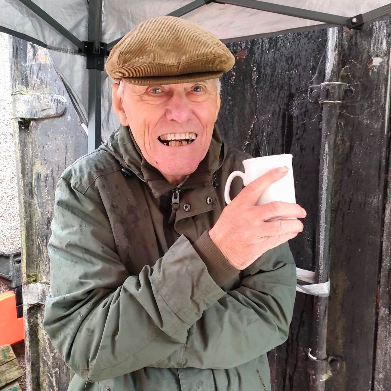 Michael, wearing green coat and brown corduroy flat hat, stands outside under a gazebo holding a hot drink and smiles for the camera