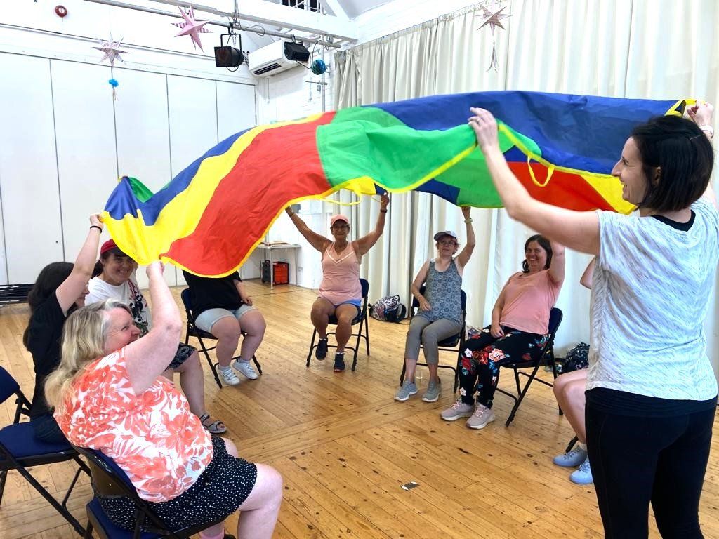 At Ashcroft Arts Centre, several FNC members are seated in a circle with arms raised holding on to a multi-coloured parachute and getting it to billow