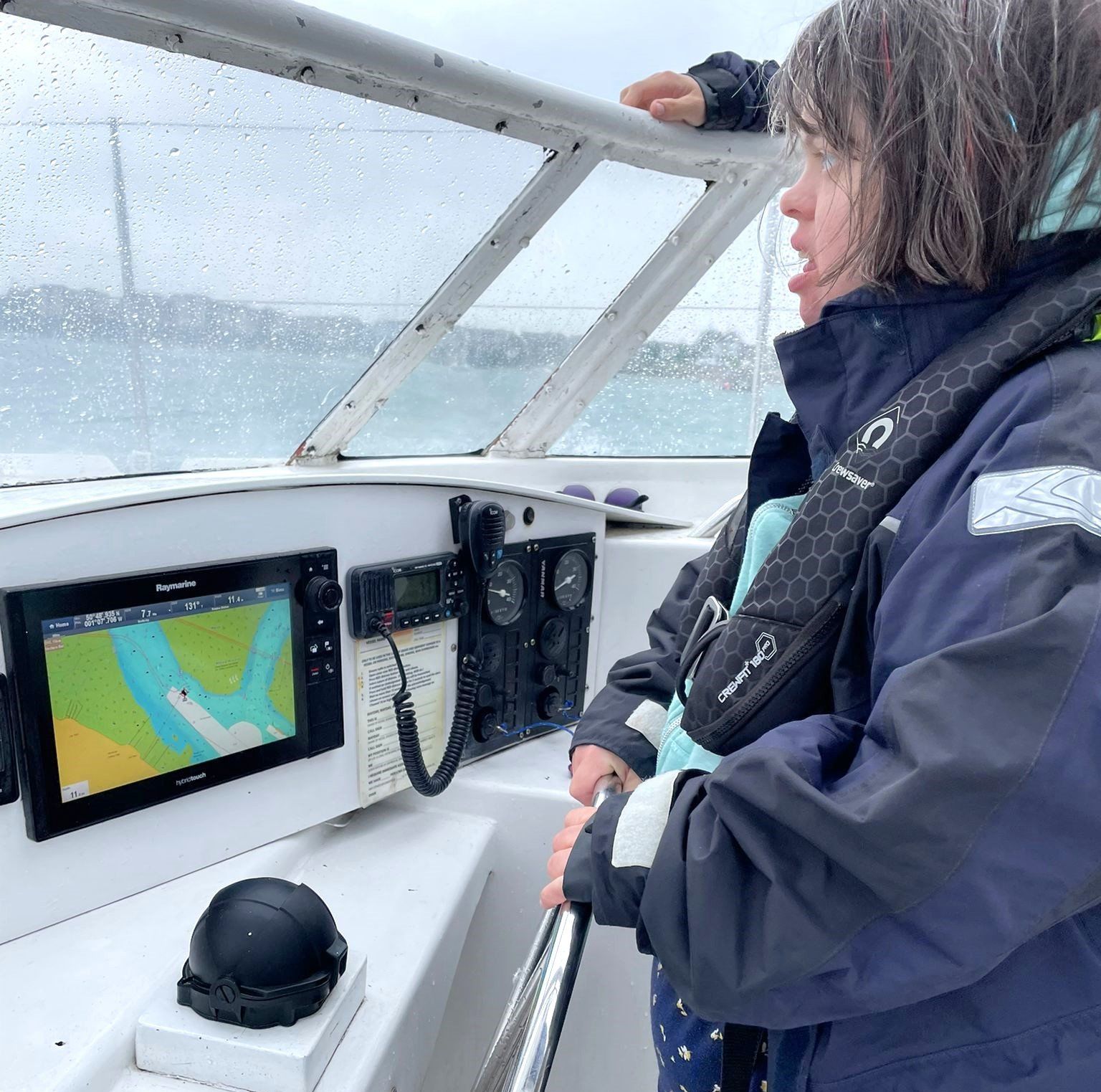 A female FNC member dressed in wet weather sailing gear and buoyancy aid  stands at the helm of the Ro Ro Project accessible catamaran in the Solent. Compass, Navigator/plotter and radio are mounted on the bulkhead in front of the helm.