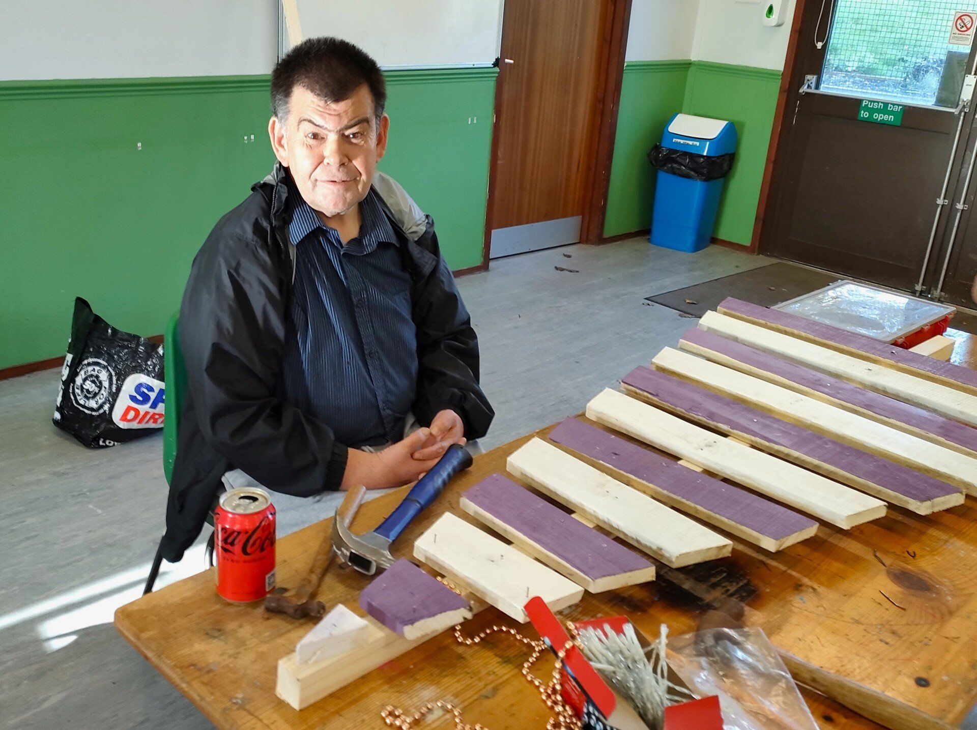 Smiling male FNC member sat at table with hammers resting on table .  Large bands of alternate purple and white painted wood nailed to central piece for a triangular tree shape that fills the table.