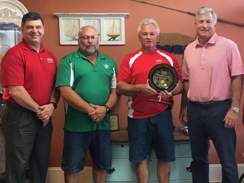 Four men stand indoors; one holds a trophy. Red, green, and pink shirts. Two wear shorts. Nautical background.