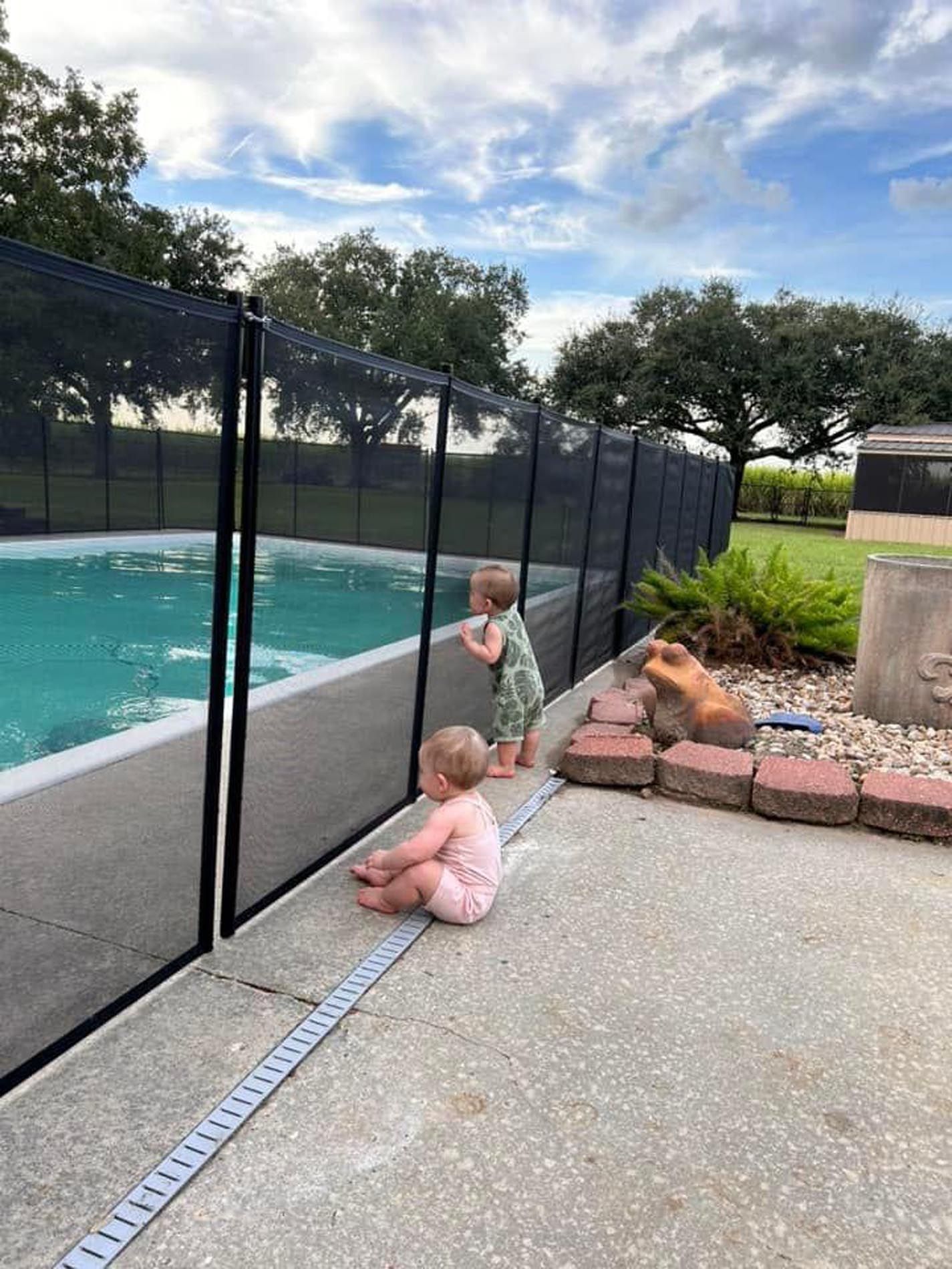 Two little girls are sitting on the side of a fence looking at a swimming pool.