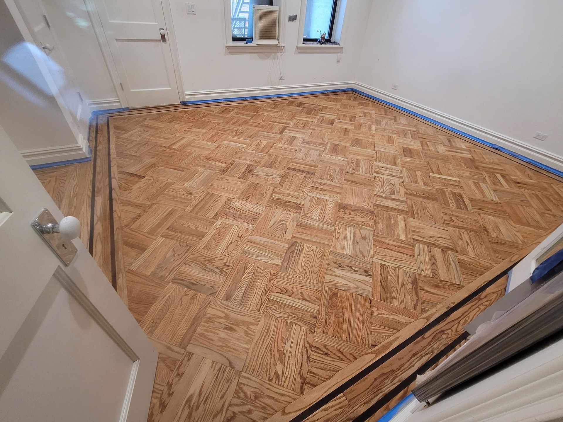 Newly installed parquet wooden flooring in a room with white walls and a doorway.
