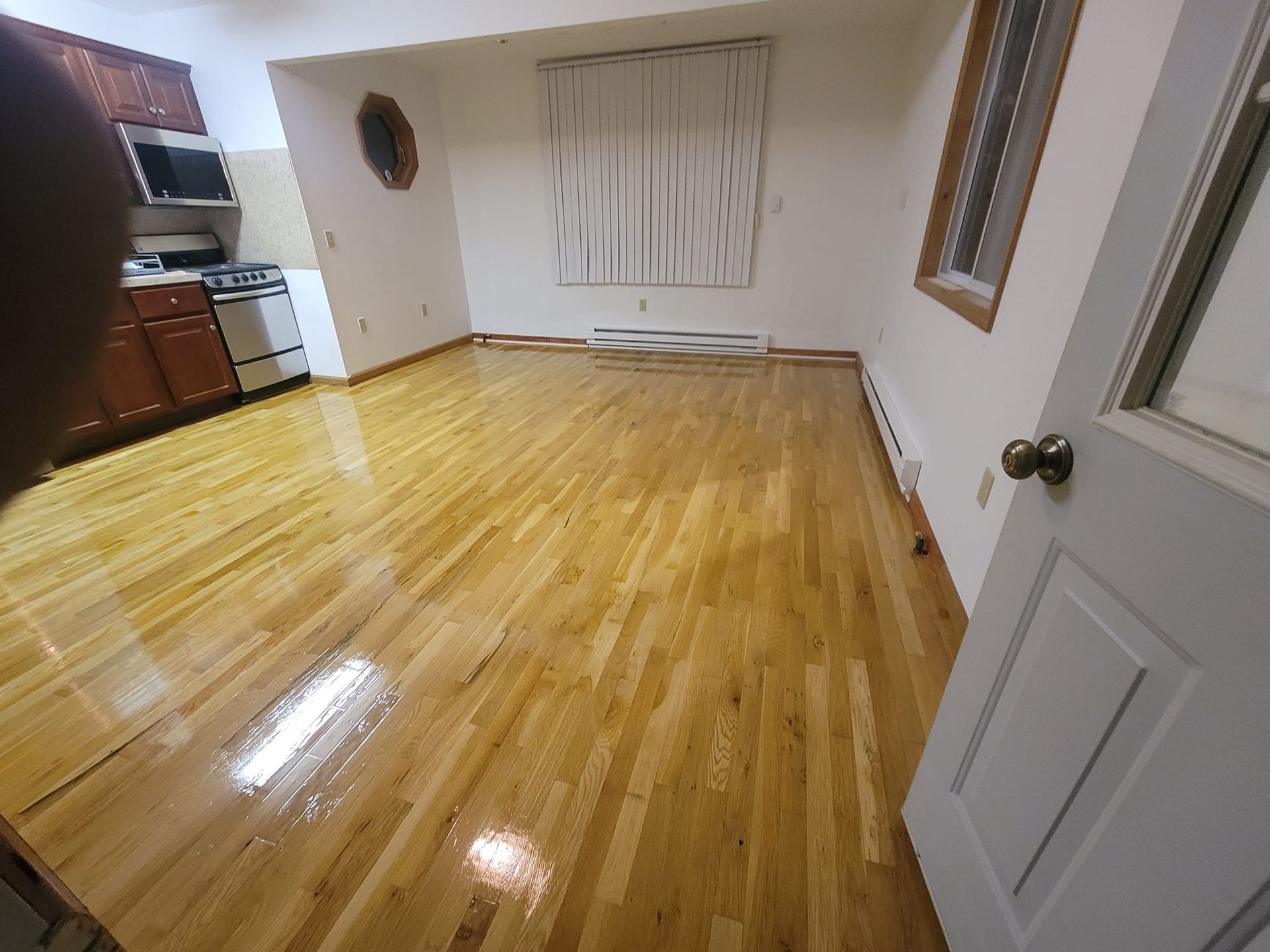 Wooden floor in a room with a kitchen in the background, a window, and a door on the right.