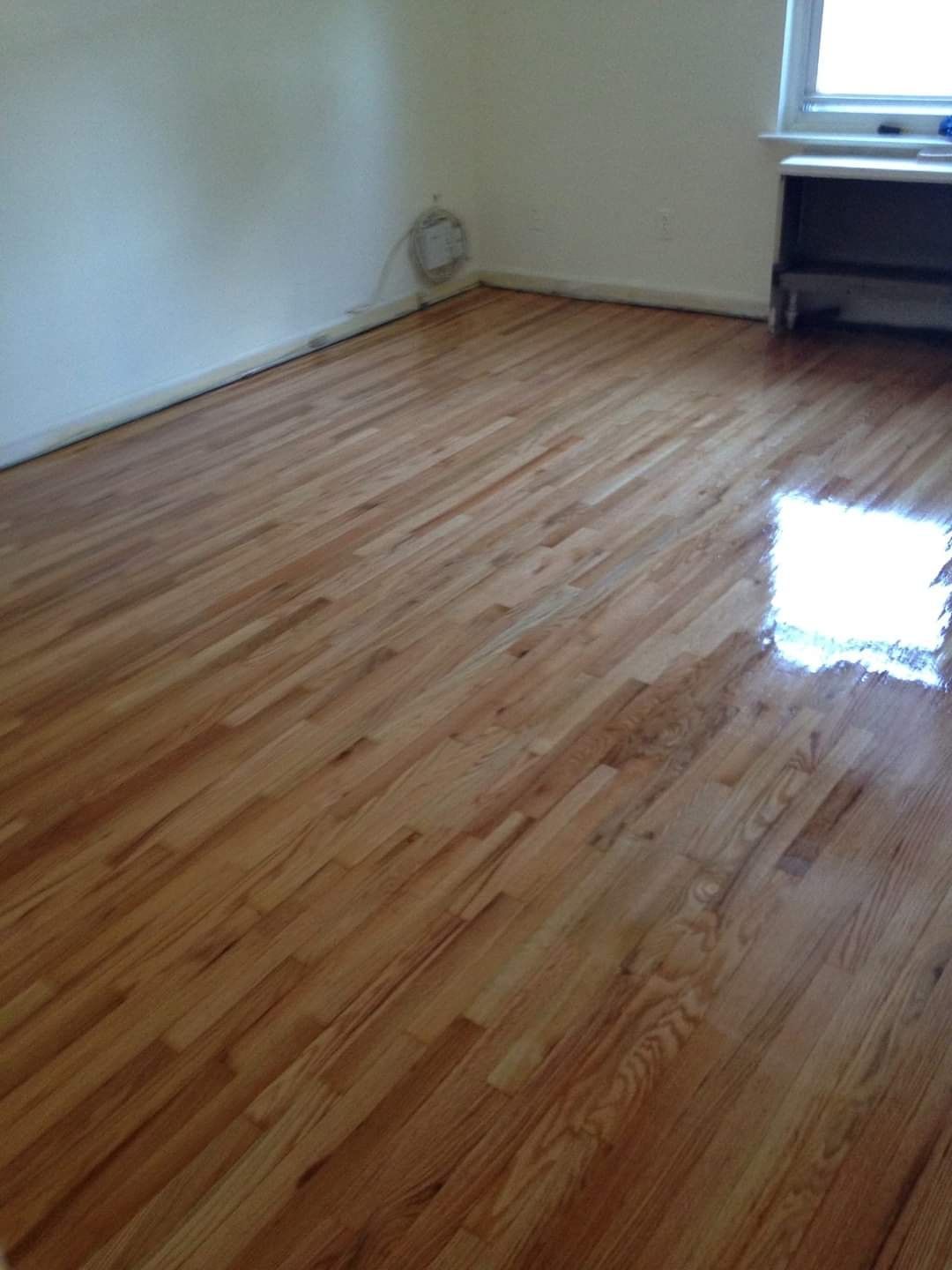 Hardwood floor in an empty room, reflecting light. Beige walls, window and cabinet visible.
