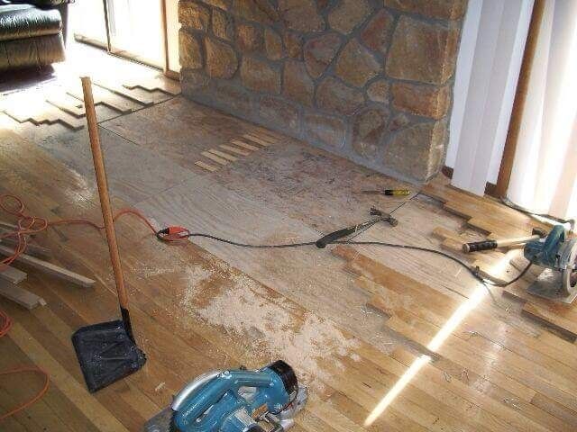 Wooden floor being repaired with scattered sawdust, tools, and a stone fireplace in the background.