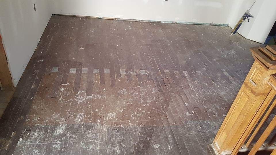 Dark wood floor with light-colored debris, a partially visible wooden staircase, and a white wall.