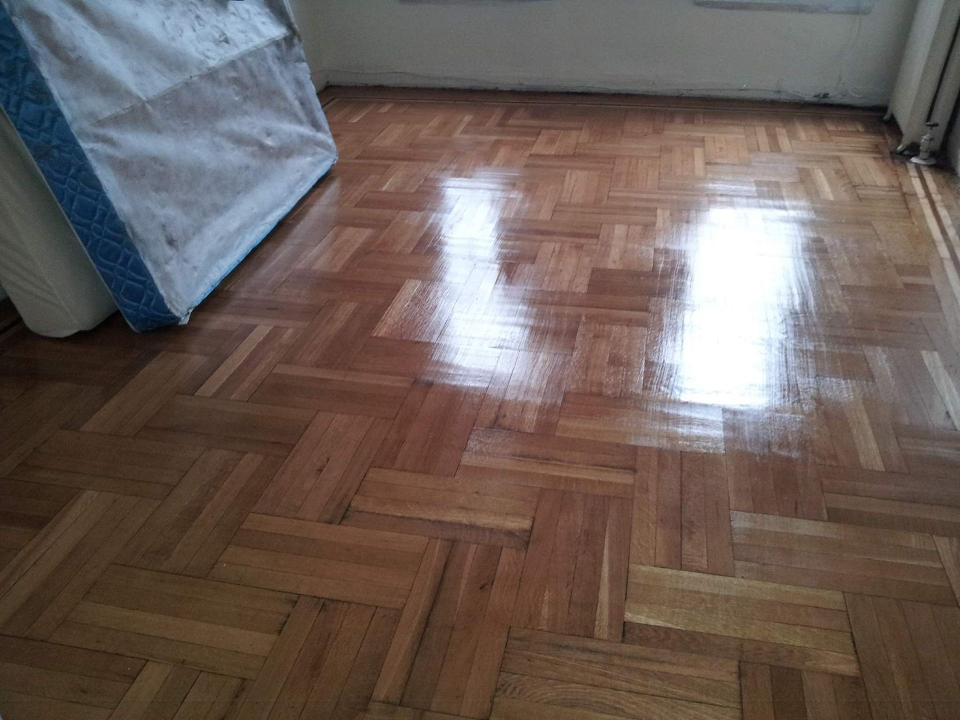 Shiny, light-brown, parquet wood floor reflecting light. A mattress leans against the wall in the corner.