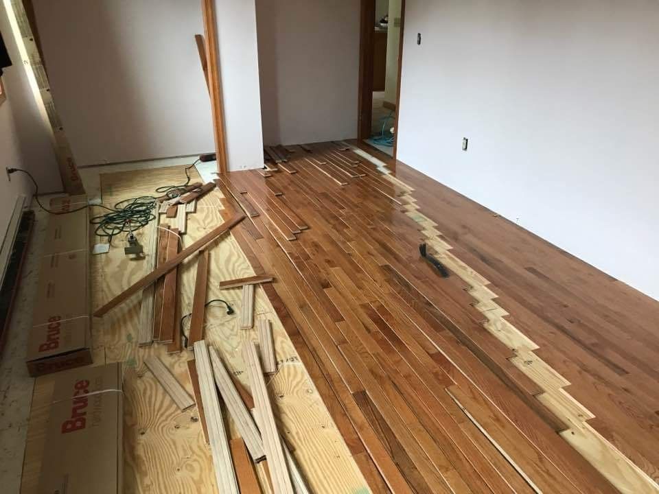 Hardwood flooring installation in progress; wood planks laid on plywood subfloor, tools and materials scattered.