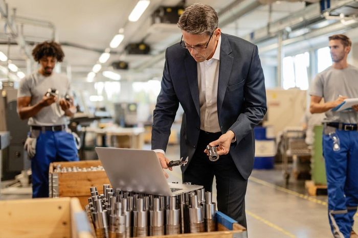 Manager inspecting metal parts on a factory floor beside two workers and a laptop cart