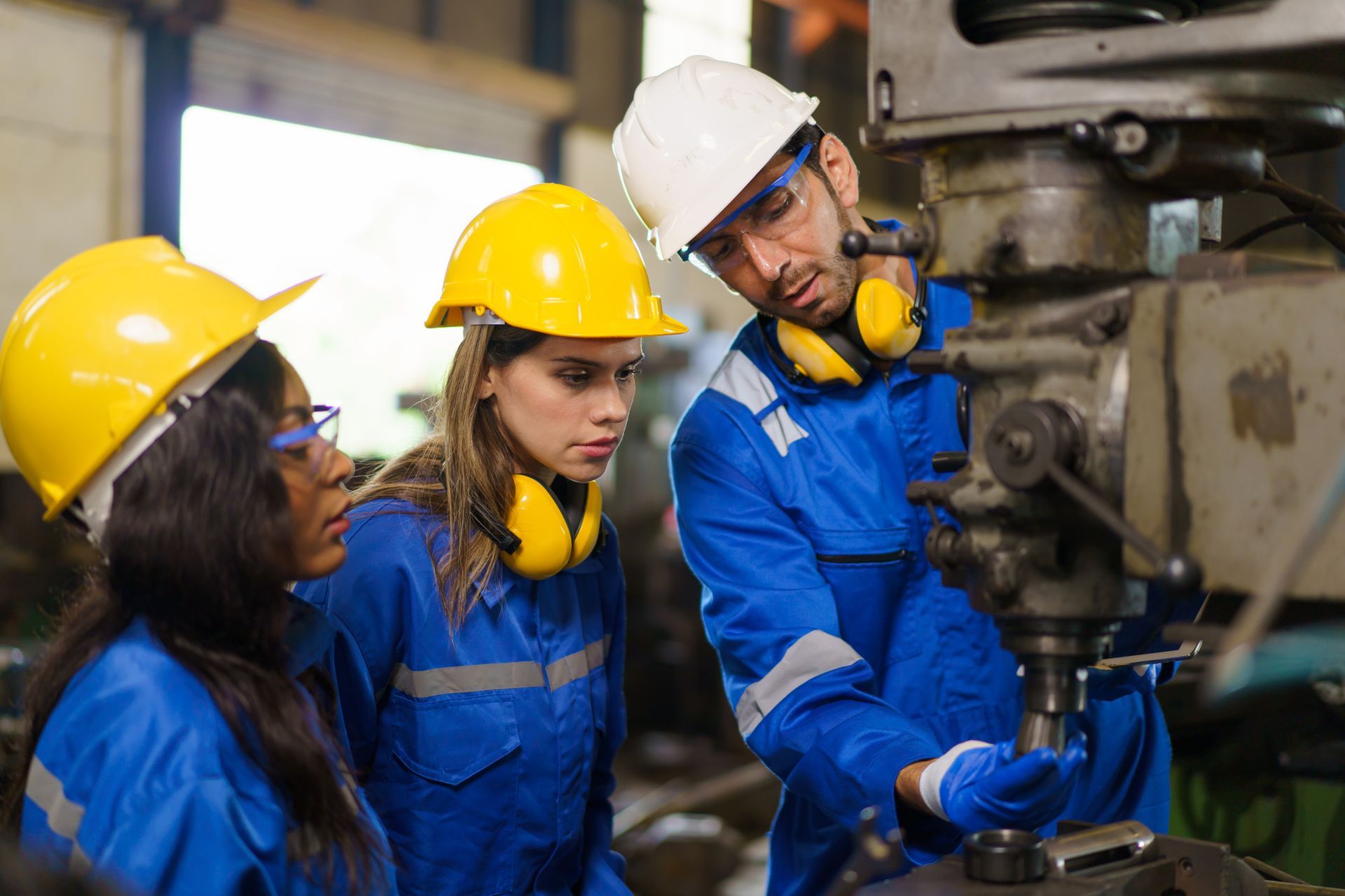 Three workers in hard hats inspect machinery in a factory, one using a tool.