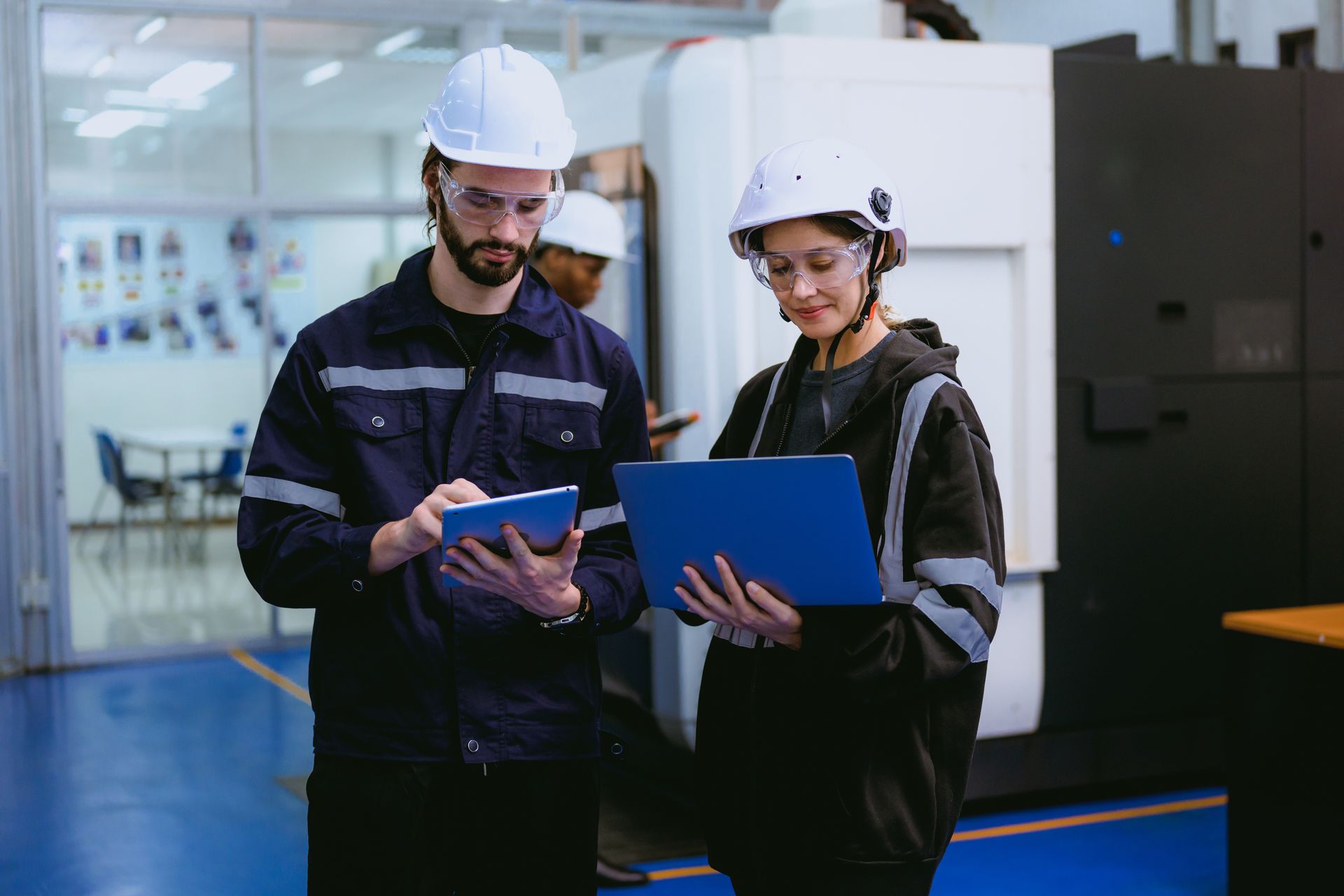 Two factory workers in hard hats reviewing a blue clipboard in an industrial facility
