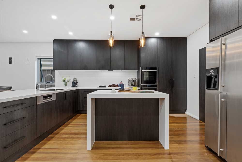 Modern kitchen renovation with dark wood-grain cabinetry, a white waterfall island, amber pendant lights, and stainless steel appliances.