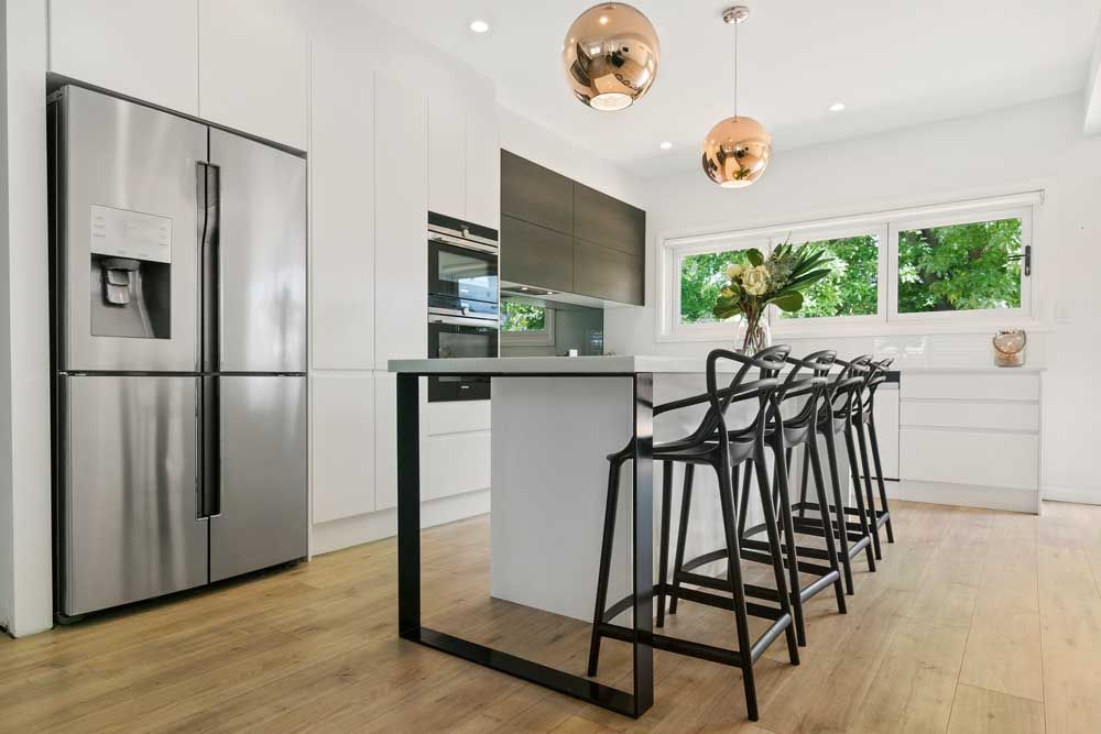 Modern kitchen renovation with a white waterfall island, black designer bar stools, copper pendant lights, and a stainless steel fridge.