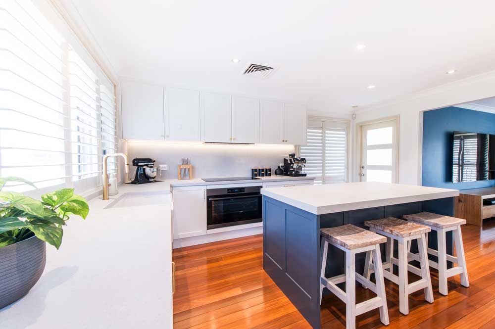 Modern kitchen renovation with navy blue shaker island, white stone benchtops, gold tap, and warm polished timber floors.