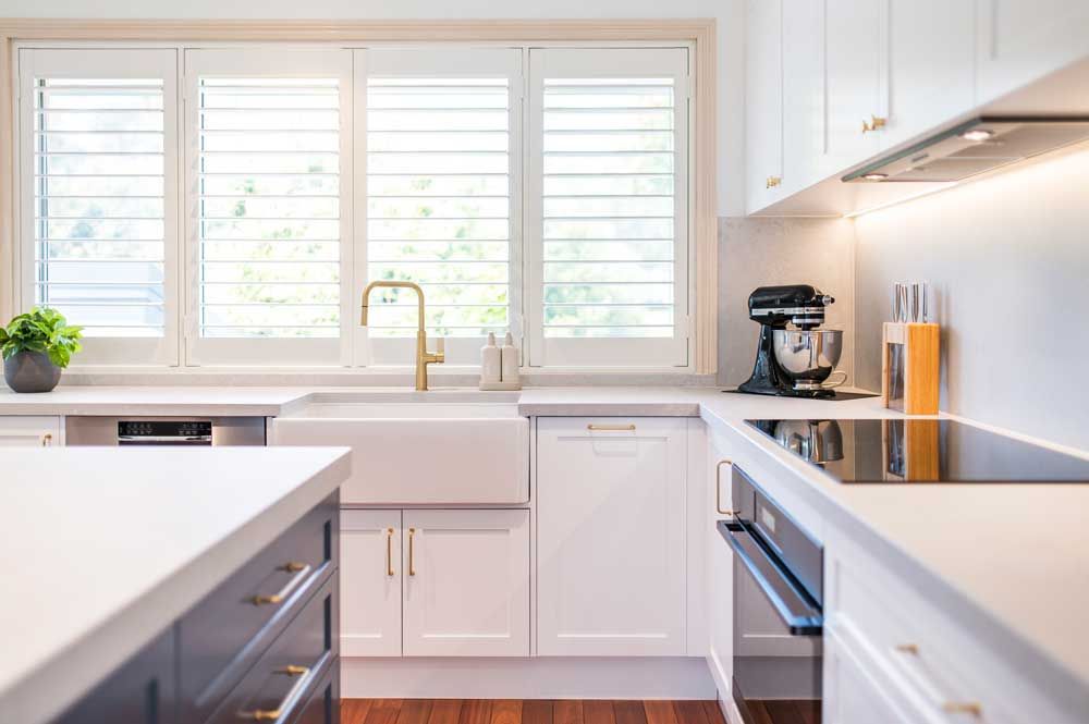 Kitchen renovation with white shaker cabinets, apron-front farmhouse sink, gold tap, and white plantation shutters.