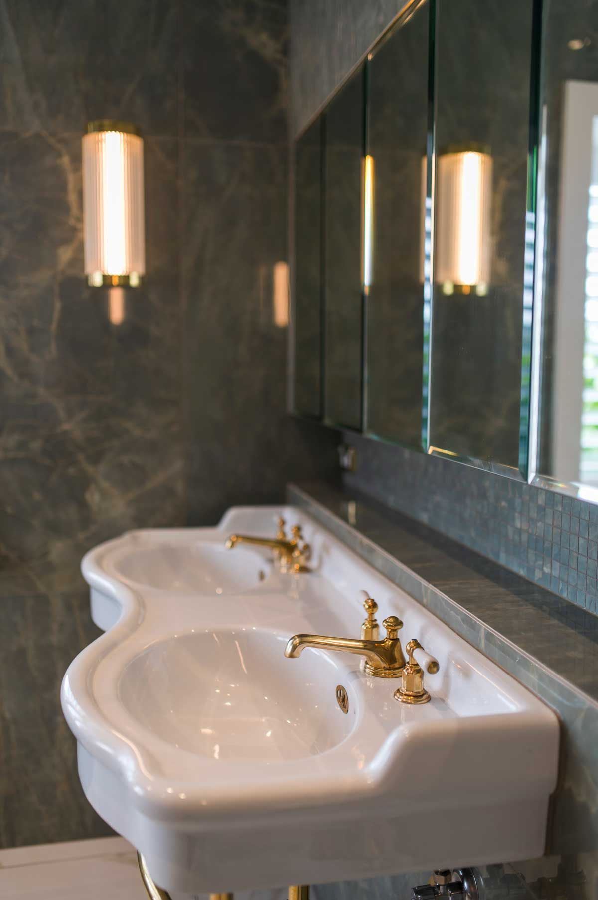 Renovation featuring a white curved double-basin sink, vintage gold taps, fluted wall sconce, and dark marble wall tiles.